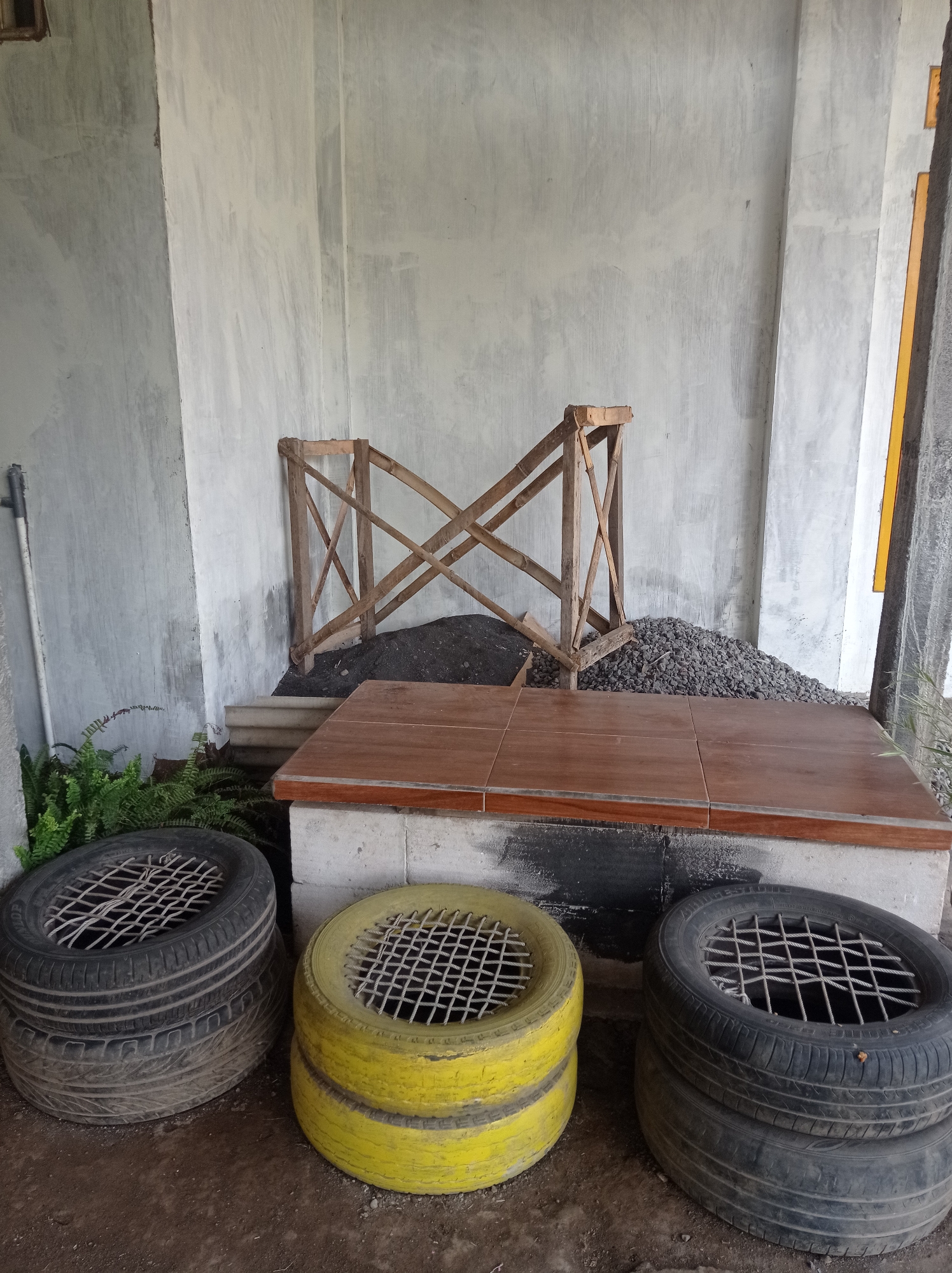 In the city of Pandanmulyo, Indonesia, an outdoor scene features a gray concrete wall with textured surfaces. In the foreground, three stacks of tires are arranged on the ground. The left stack consists of two black tires, the middle stack has two yellow tires, and the right stack is made of two black tires. Each tire has a metal grate placed inside its opening. Behind the tires, a raised platform is constructed from concrete blocks topped with wooden planks. To the left of the platform, a cluster of green ferns grows. Behind the platform, there is a pile of dark soil and a heap of gravel. A wooden structure, resembling a fence or support, is positioned against the wall behind the gravel pile. A metal pipe is visible on the far left, attached to the wall. The overall environment suggests an unfinished outdoor area or a repurposed space.