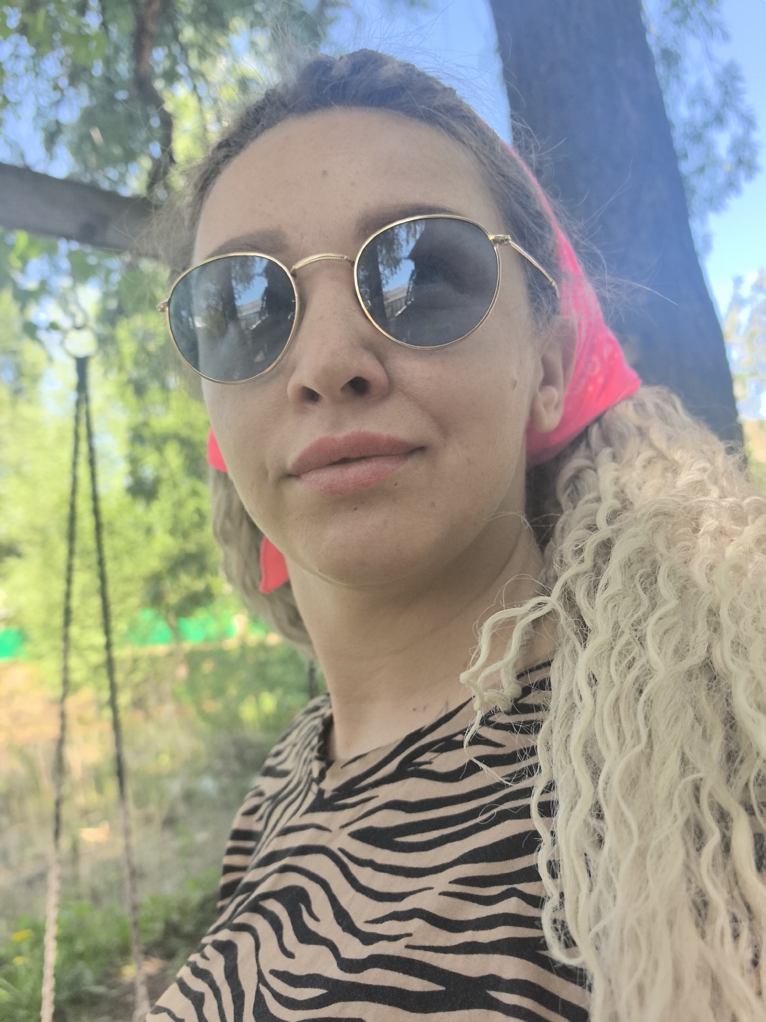 A medium shot captures a woman outdoors on a sunny day. She is wearing round, gold-framed sunglasses, a pink bandana, and a zebra-striped shirt. Her hair is blonde and curly. The background features greenery, a tree trunk, and what appears to be a swing set. There is no evidence of any specific city location, and the city of Noviy, Russia cannot be confirmed from this image.
