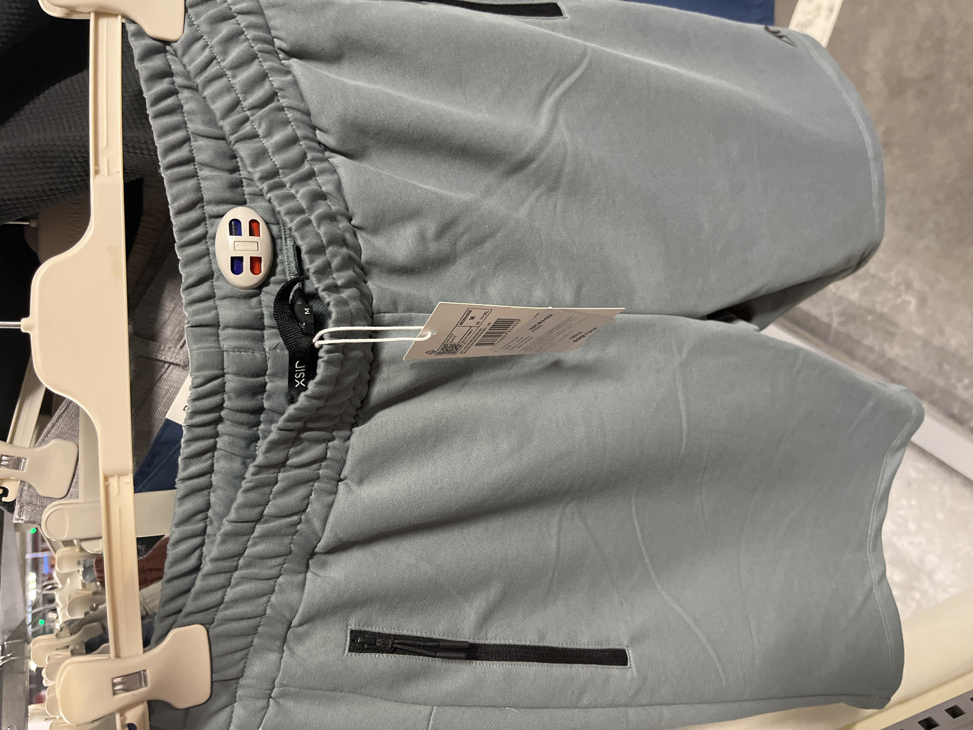 This media file is a close-up shot of a pair of light blue-grey sweatpants hanging on a clothing rack. The sweatpants have an elastic waistband and zippered pockets. A small white tag with red and blue accents is attached to the waistband. Another white tag, with visible text including a barcode and sizing information, is hanging from a string on the sweatpants. The background shows other clothing items on hangers and a blurred store environment. The image appears to have been taken indoors, likely in a retail setting, given the clothing racks and the general ambiance. No people are visible in the frame. The lighting suggests it is daytime. The text on the visible tag is mostly illegible due to the angle and resolution, but "M" is clearly visible indicating the size. The overall impression is a product shot focusing on the details of the garment.