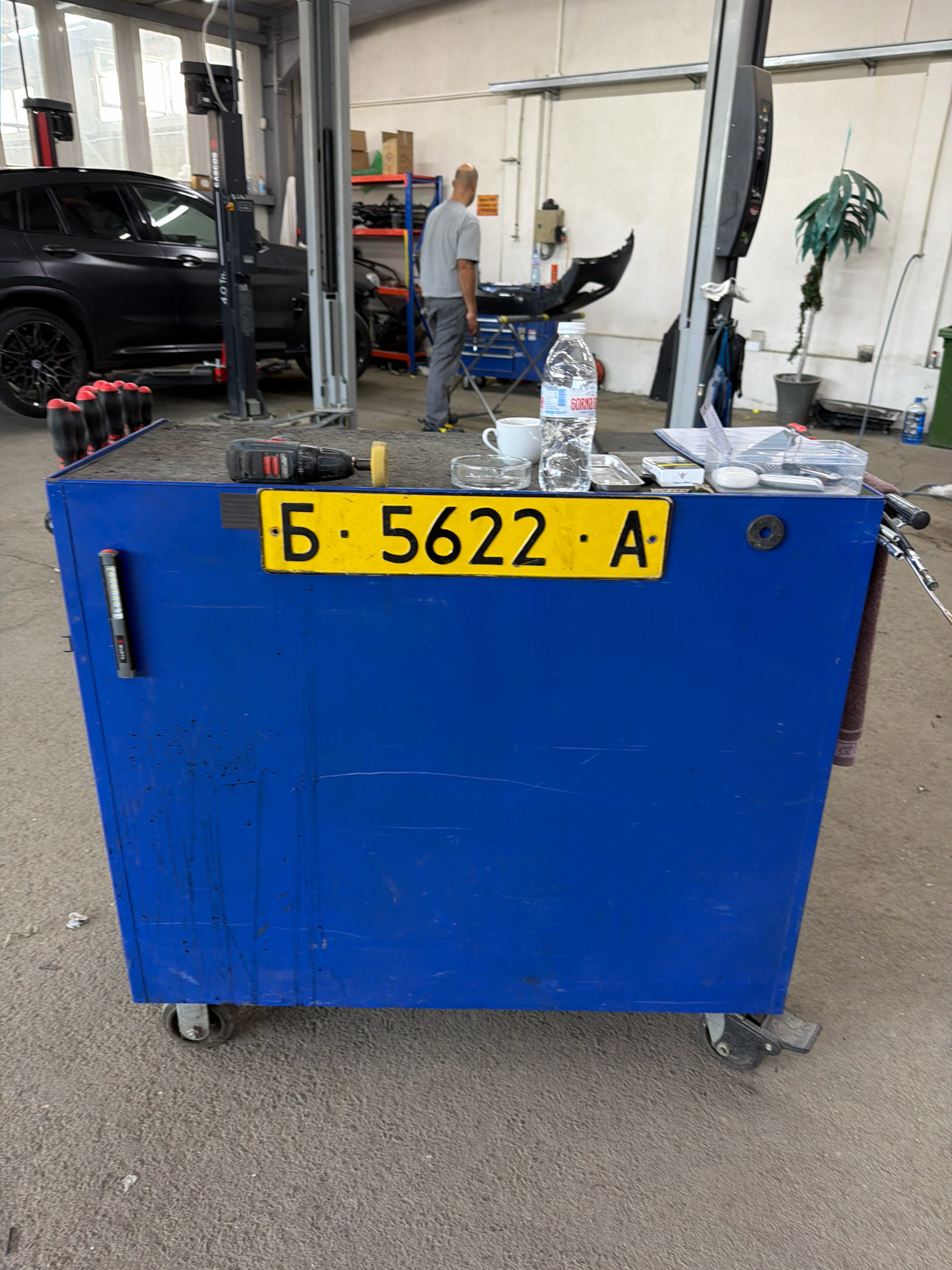 The image depicts a blue metal toolbox with wheels, situated in what appears to be a car repair workshop in Burgas, Bulgaria. A yellow license plate with the characters "Б 5622 А" is affixed to the front of the toolbox. On top of the toolbox, several items are visible, including a cordless drill, a white ceramic mug, a plastic bottle of water, a small metal tray, a pack of cigarettes, and a clear plastic container holding various small objects. A red LED flashlight is attached to the left side of the toolbox. In the background, a black car is parked next to a vehicle lift. A man in a grey t-shirt and pants is standing near the car, facing away from the camera. Shelving units with various items are also visible in the background. The floor is concrete, and the overall environment suggests a working garage.