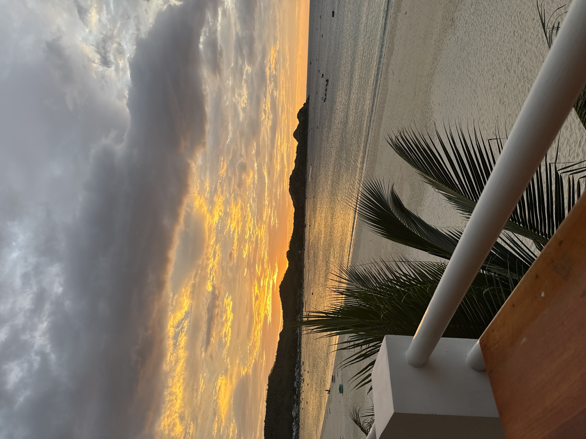 The image depicts a scenic view of Bai Xep beach in Song Cau, Vietnam, during what appears to be sunset. The sky is dramatic, with heavy gray clouds interspersed with vibrant streaks of orange and yellow from the setting sun. Below the sky, a calm sea reflects the warm hues of the sunset. The coastline is lined with lush green hills, and a small village can be seen nestled along the shore. The sandy beach is largely empty, with a few scattered boats visible in the water. In the foreground, a white railing and a wooden surface suggest a vantage point, possibly a balcony or terrace, from which the photograph was taken. Palm fronds are visible, adding to the tropical ambiance. The overall scene is tranquil and picturesque, capturing the beauty of a coastal sunset.