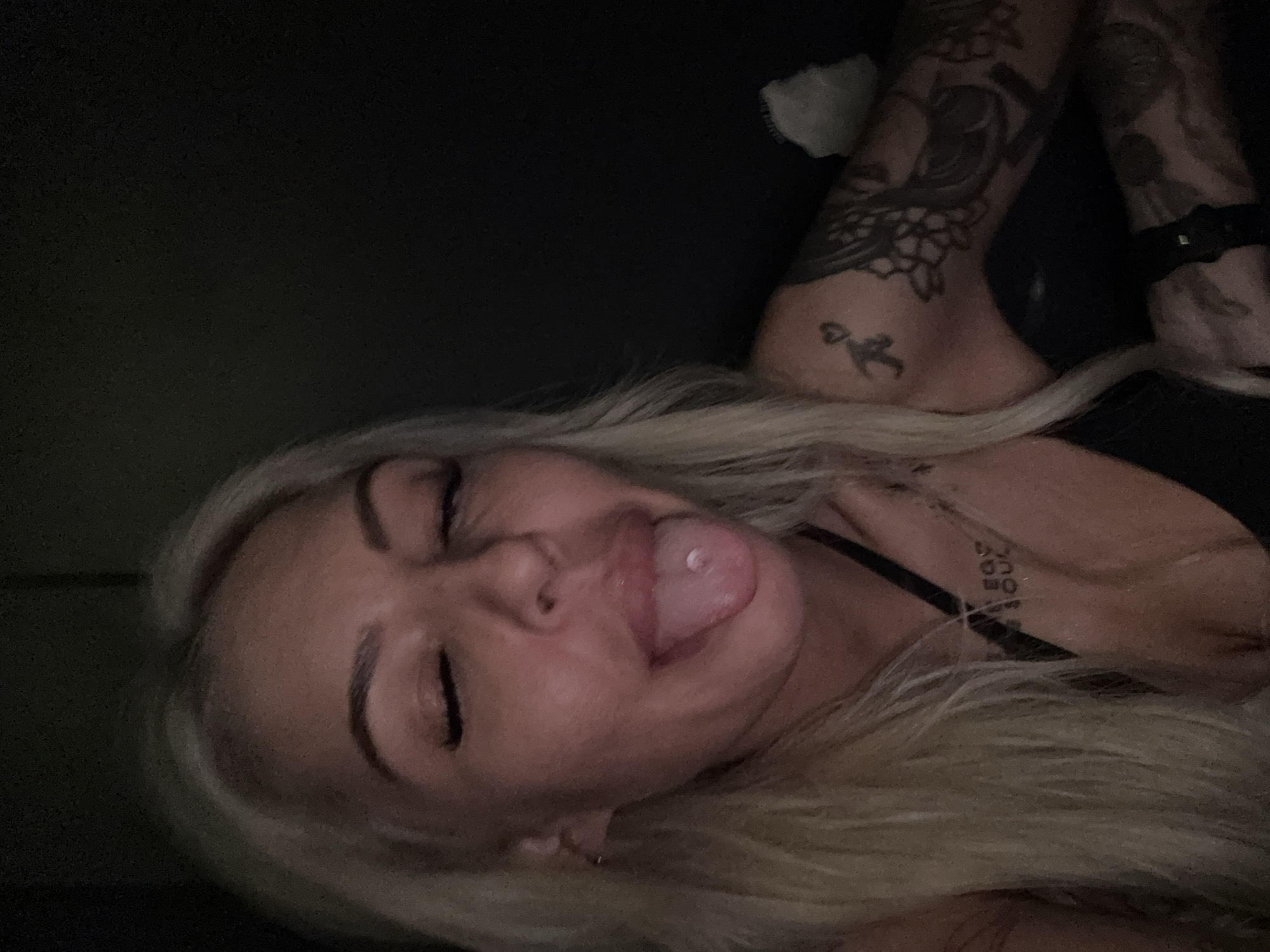 A fair-skinned woman with long blonde hair is lying down, eyes closed, sticking out her tongue. The woman has dark eyebrows, a nose ring, and tattoos on her arm and chest. The background is dark and indistinct. There is no indication of the location being Sofia, Bulgaria.