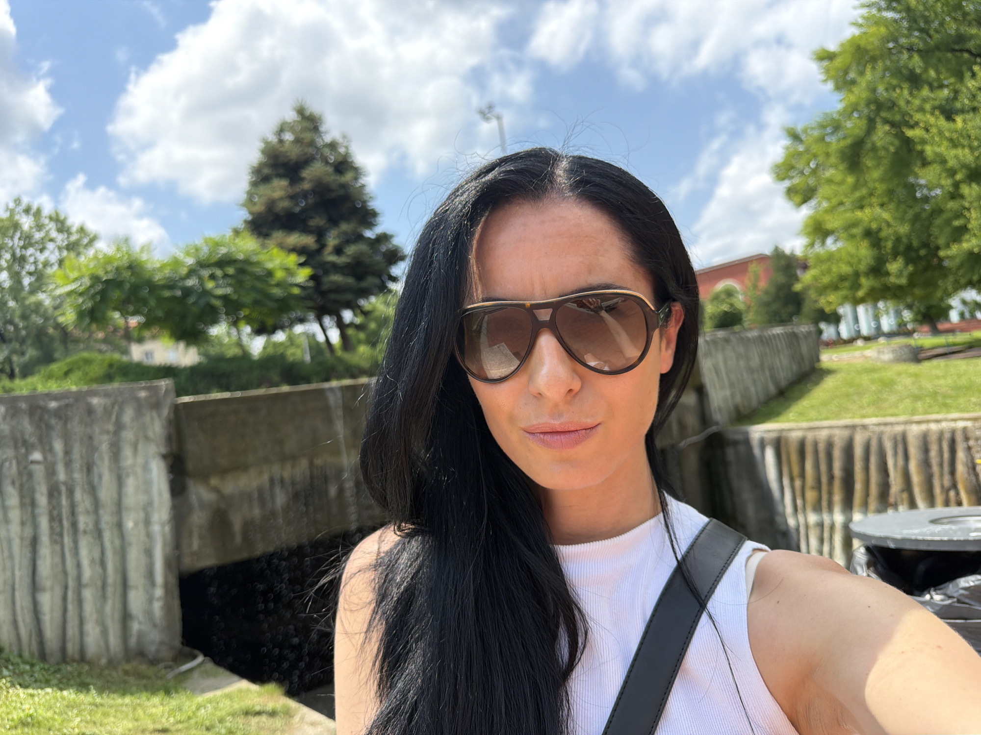 This is a sunlit selfie of a woman taken outdoors in a park or public green space. The woman, an adult with long, dark hair parted down the middle, is positioned slightly to the left of the frame, looking directly at the camera. She is wearing large, dark aviator-style sunglasses with a bronze-toned frame, obscuring most of her eyes, and a white ribbed sleeveless top. A black shoulder strap from a bag runs across her chest and right shoulder. Her right arm, holding the camera, is visible in the bottom right corner of the frame. Her expression is neutral.
The background features a clear blue sky with scattered white cumulus clouds, suggesting a bright daytime setting. Below the sky, there are concrete retaining walls, possibly for a canal or water feature, which show texture and weathering. Beyond these structures, a vibrant green landscape unfolds with areas of grass and numerous trees. Prominent trees include a tall, dark evergreen behind the woman's head on the left, and a lush, light green deciduous tree on the right. In the distance, indistinct buildings with reddish roofs are visible through the foliage. A dark grey public trash can is partially visible on the far right. The scene is located in Pleven, Bulgaria. No text is visible in the image.