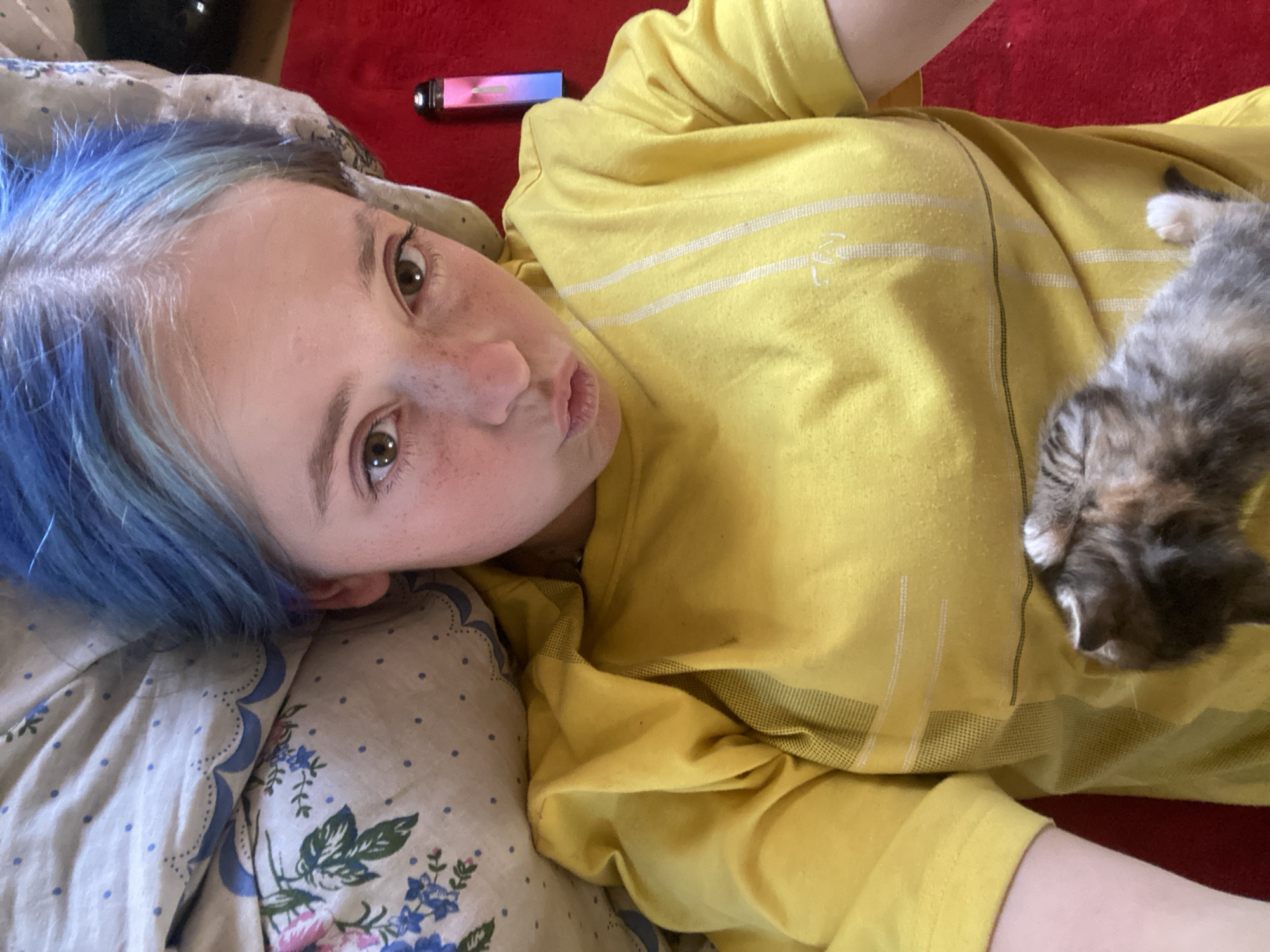 A close-up, low-angle shot shows a person with bright blue hair lying on their side, looking at the camera. They have fair skin with freckles and are puckering their lips. The person is wearing a bright yellow t-shirt with white stripes and a logo. A small, fluffy kitten with grey, brown, and white fur is nestled on the person's chest. The person is lying on a patterned blanket with blue and white floral designs, and to the right, a red surface is visible, possibly a carpet or bedspread. In the background, near the top left corner, a rectangular object with pink and blue light is visible, possibly an electronic device. The lighting suggests daytime indoors. The overall mood appears relaxed and affectionate due to the presence of the kitten. There is no visible text.