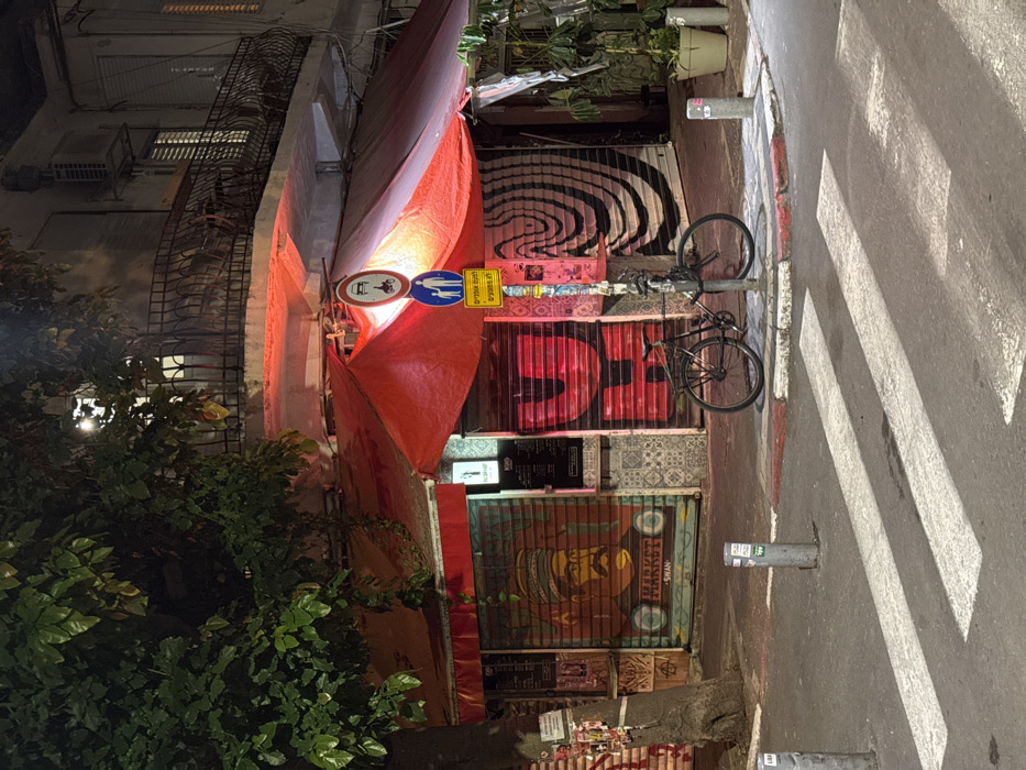 A narrow street scene at night features a building adorned with graffiti-covered roll-down doors. A red awning extends from the building over the sidewalk. Street signs on a pole near the awning depict pedestrian and stroller crossing. A bicycle is locked to the same pole. The street has a crosswalk. Green vegetation frames the left side of the image, while the building facade is visible on the upper left.