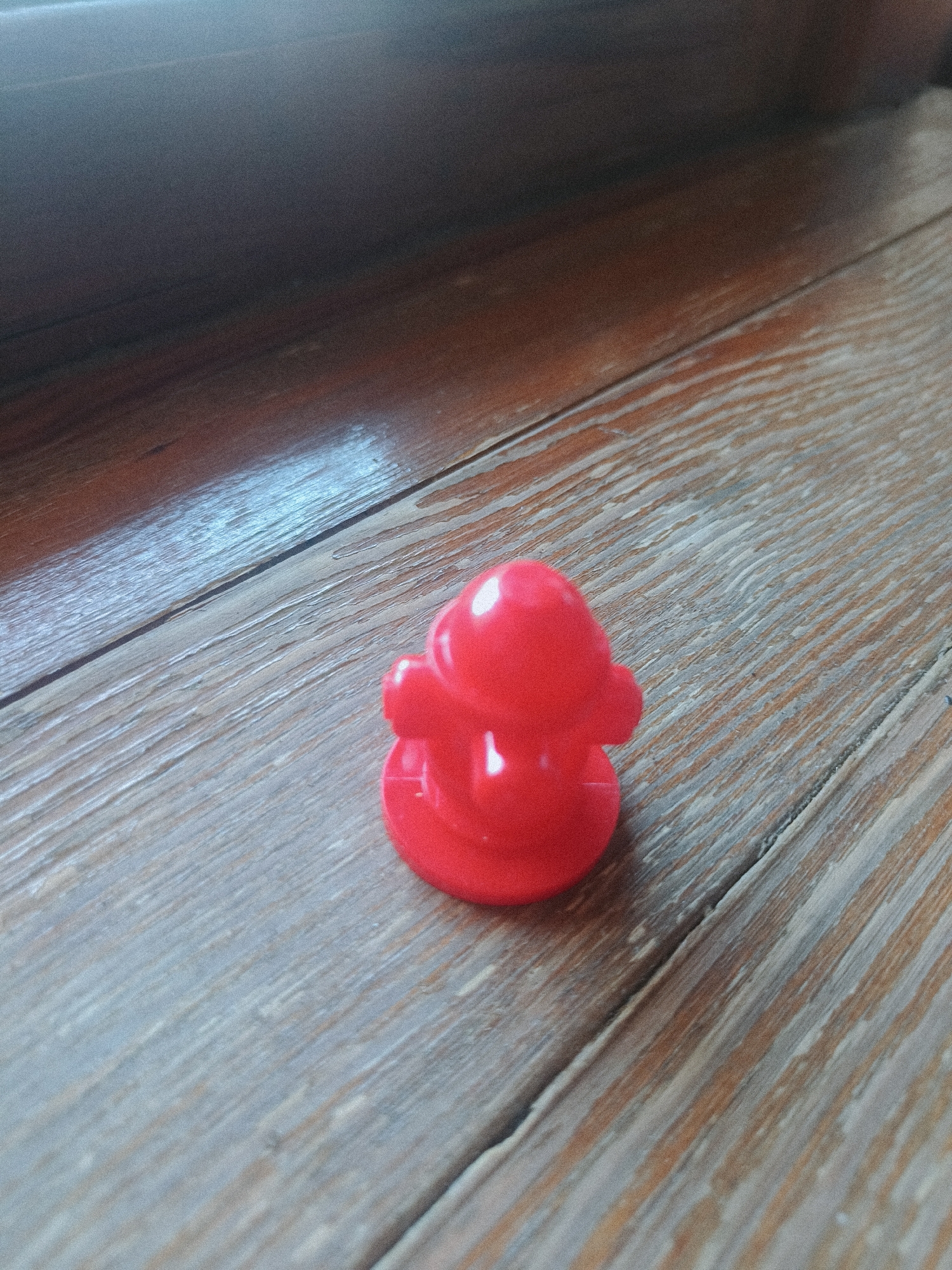 A red, plastic duck figurine sits on a weathered wooden surface. The duck is small and appears to be a bath toy. The wooden surface has visible grain and appears to be made of planks. The lighting suggests an indoor environment, with a shadow cast from an unseen object. No specific location in the city of Odesa, Ukraine can be determined from the image.
