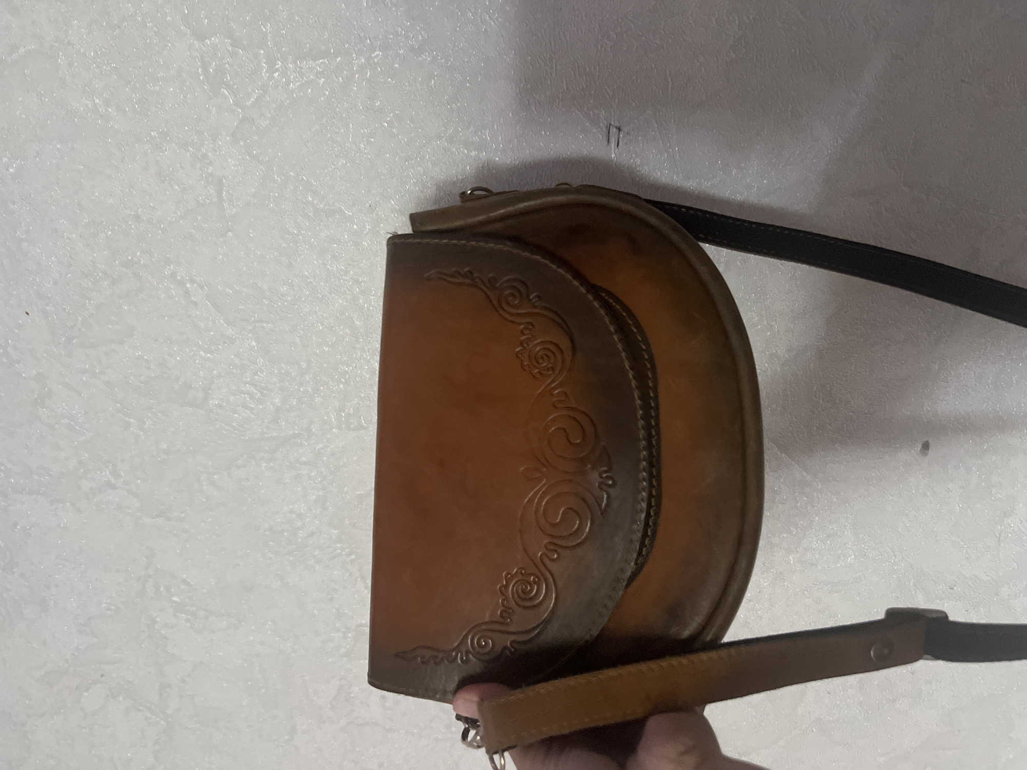 A person's hand is holding a brown leather crossbody bag. The bag has a flap closure with ornate embossed designs and a dark strap. The bag is positioned against a textured white wall, possibly in an indoor setting. The overall lighting suggests a dimly lit environment. There is no indication of the specific location being Şamaldı-Say, Kyrgyzstan.