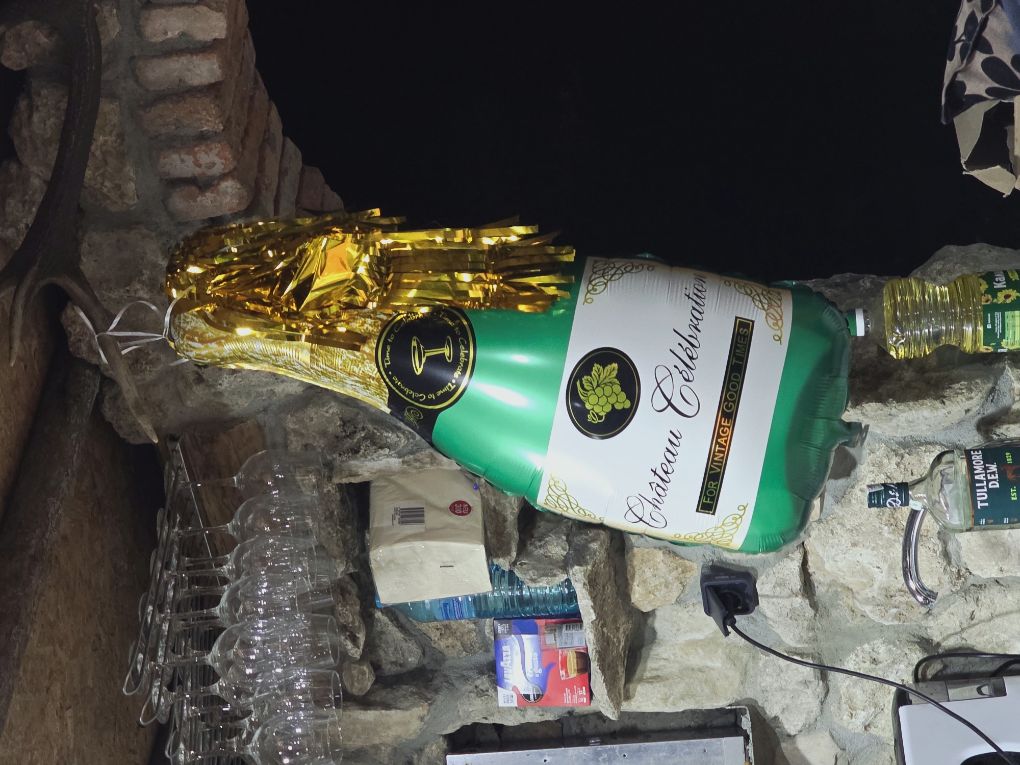 A champagne bottle-shaped balloon with a gold foil fringe hangs from the ceiling in a dimly lit room. The balloon features a label that reads "Château Célébration For Vintage Good Times." Beneath the balloon, on a stone shelf, are various items including a bottle of Tullamore D.E.W. Irish whiskey, a bottle of vegetable oil, and a Lavazza coffee product. Wine glasses are visible hanging from a rack above. The walls are constructed of rough stone. The scene appears to be in a bar or a home with a rustic aesthetic, potentially in Varna, Bulgaria, given the context. There are no people in the image.