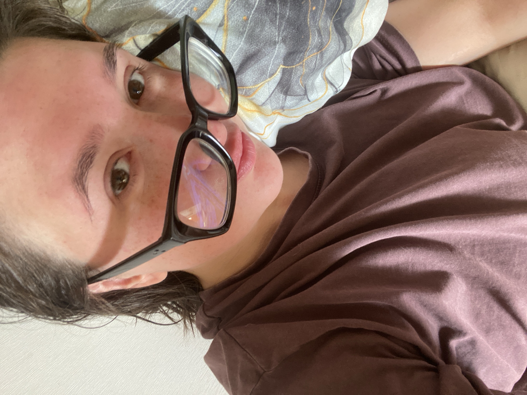 The image shows a close-up, selfie-style view of a young person lying down. They are wearing black-rimmed glasses and a dark purple t-shirt. Their hair is dark and appears slightly damp. The person is looking towards the camera, and their head is resting on a pillow with a patterned cover featuring gray, white, and orange wavy lines. The background to the left is a plain white wall. The overall lighting is soft and appears to be indoor lighting. There is no discernible information in the image to indicate a specific location within the city of Chaikovsky, Russia.