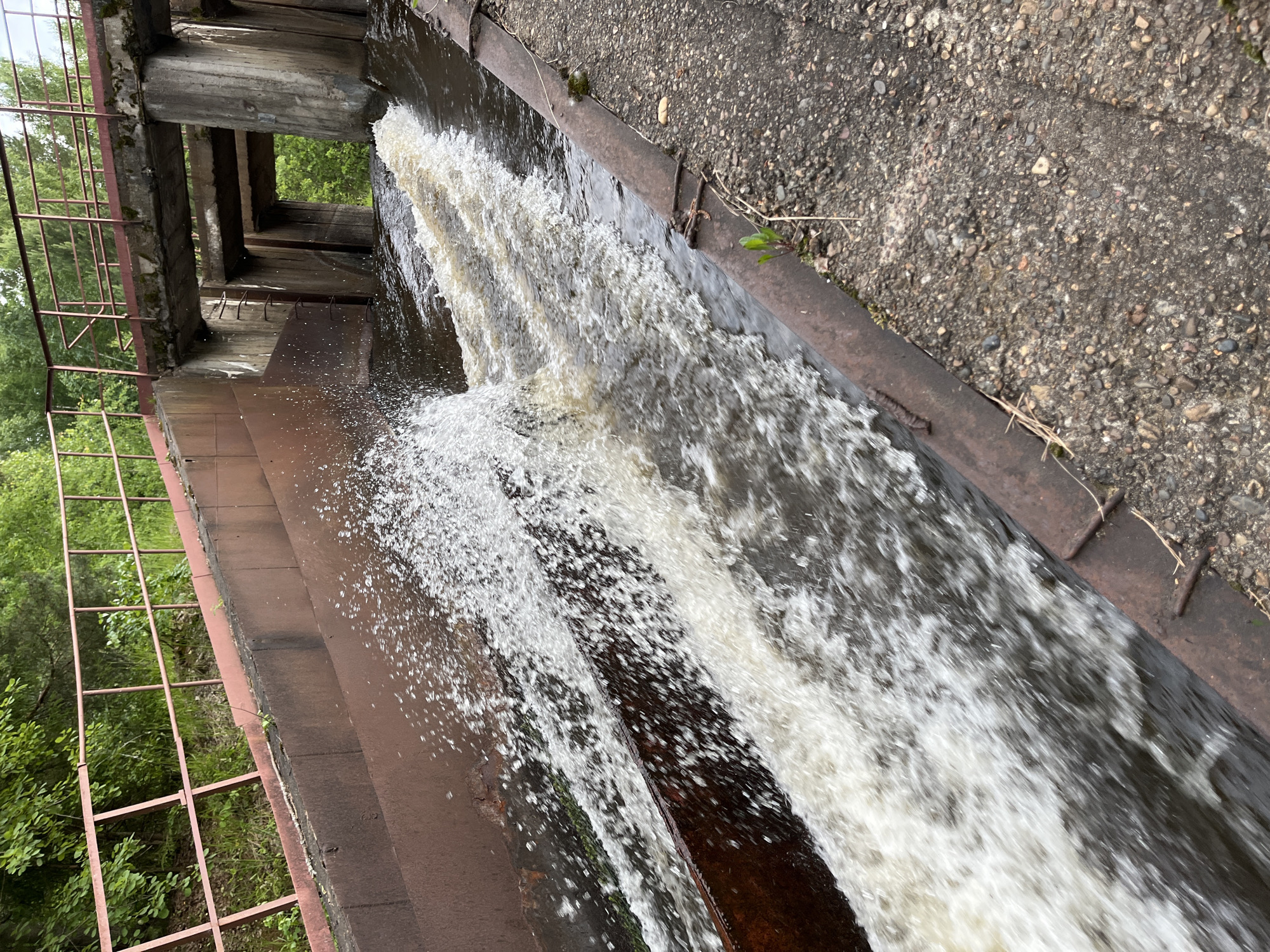 The image shows a spillway with water flowing through it. The spillway is constructed of concrete and rusted metal. There is a metal railing on the left side of the spillway, and trees and foliage are visible in the background. The water is turbulent and appears to be flowing rapidly. The scene is outdoors, and the weather appears to be overcast. The location is identified as Chernushka, Russia.