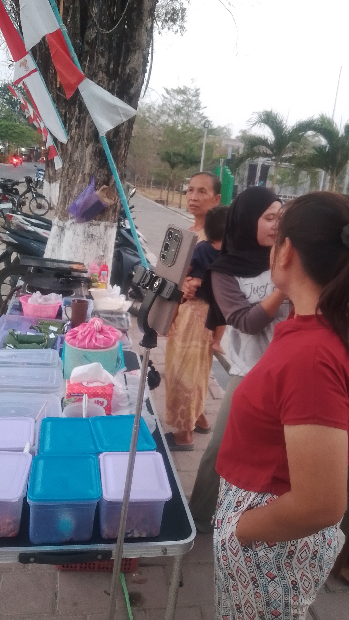 The image shows a street scene in Karanglangu, Indonesia, featuring a food stall with various food items in plastic containers. A woman in a red shirt and patterned pants stands at the front of the stall, with her hand in her pocket. Behind her, a woman in a hijab is holding a baby, and another older woman stands behind them. A selfie stick with a phone is mounted on a stand in front of the stall. In the background, there are trees, palm trees, and some motorcycles. Red and white flags are displayed on a nearby tree.