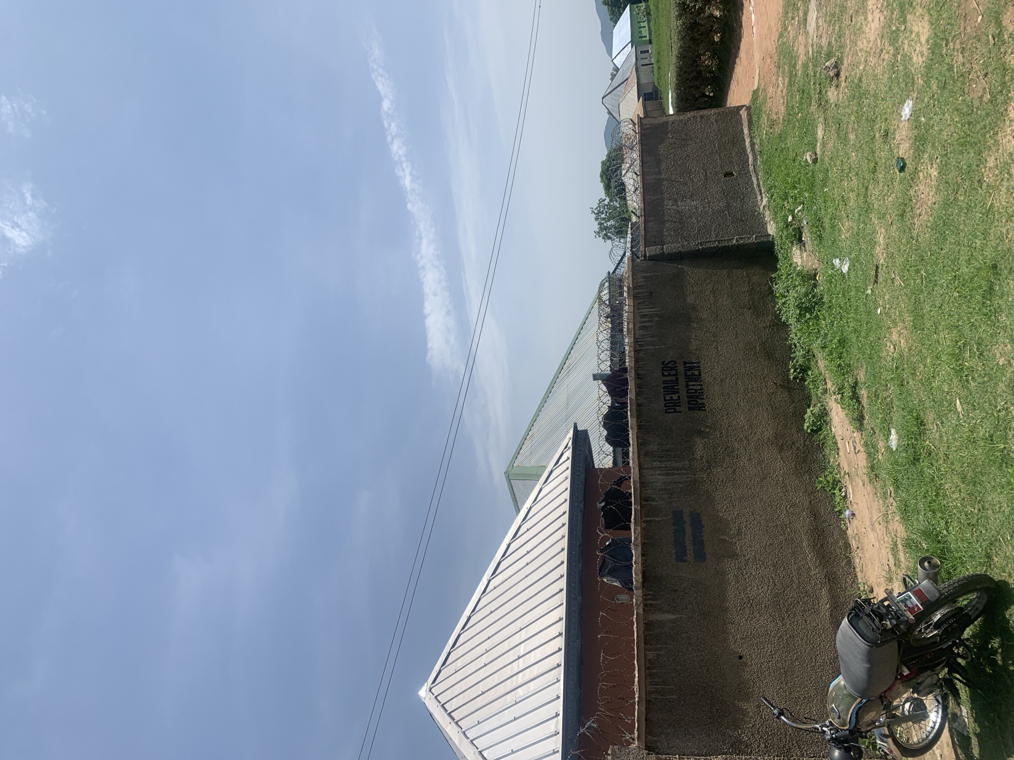 In Jauro Yinu, Nigeria, an outdoor scene depicts a gray concrete wall topped with barbed wire. On the wall, the words "PREVAILE" and "APARTMENT" are painted in black. To the left of the wall, a silver-roofed building with red walls is visible, also featuring barbed wire on its top. Behind this building, a green-roofed structure is partially visible. A motorcycle with a gray seat and black accents is parked on the grassy ground to the left of the wall. The sky is blue with scattered white clouds. In the background, several houses with different colored roofs are situated behind a neatly trimmed green hedge, with trees and hills in the distance. The ground is covered with grass, dirt, and some scattered litter.
