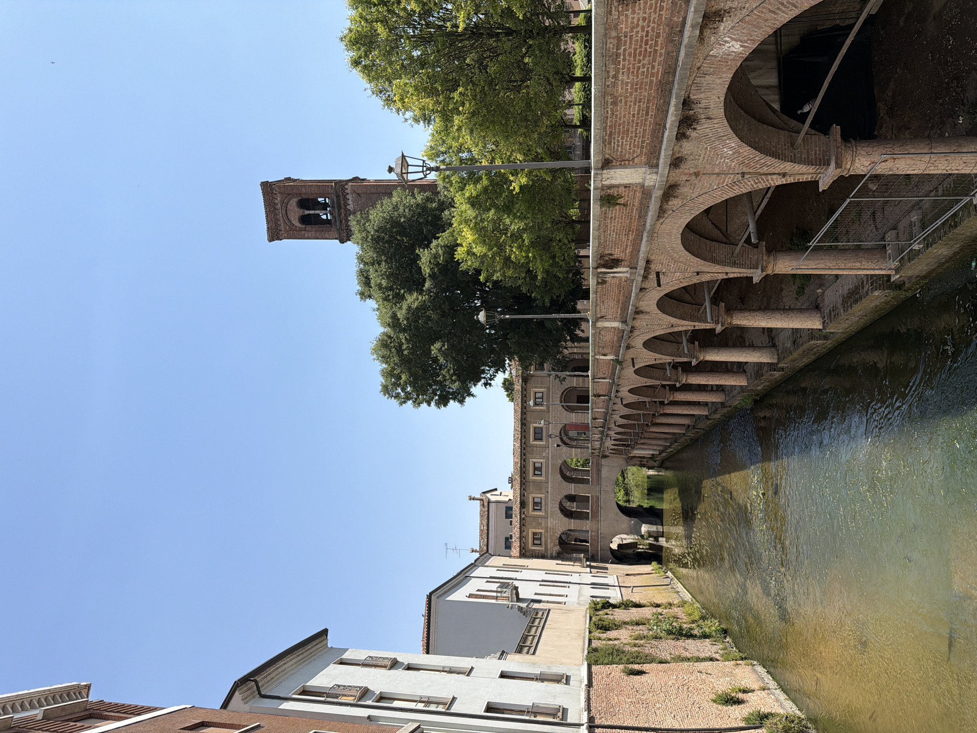 This image captures a tranquil canal scene in Mantua, Italy, distinguished by its unique architectural elements. The water, tinged with a brownish-green hue, flows gently through the canal. Overlooking the water is an arched arcade structure made of red brick, supported by sturdy pillars. Adjacent to the canal and arcade is a row of buildings, varying in height and architectural style. Some buildings have white facades with windows, while others feature brick exteriors. A prominent bell tower rises in the background, its brick construction mirroring the arcade. Lush green trees and vegetation add a natural element to the urban landscape. The sky above is clear blue, suggesting a sunny day. Overall, the scene combines architectural history with the natural beauty of the waterways, creating a peaceful snapshot of Mantua.