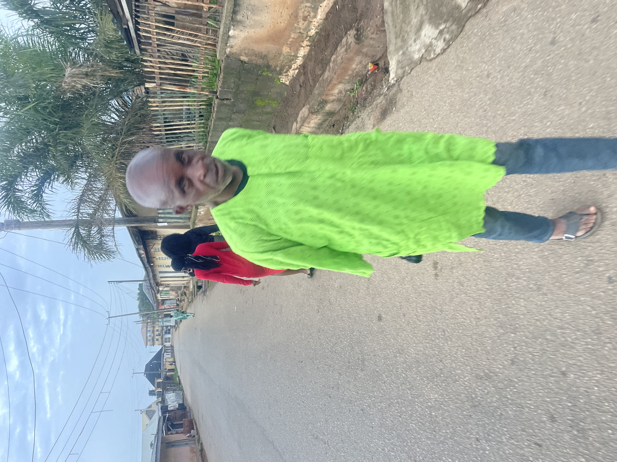 In Jos, Nigeria, on a paved street, a man in a lime green traditional outfit and blue jeans walks toward the camera. His head is shaved, and he has a prominent brow bone. Behind him, a woman in a red dress walks away from the camera, followed by another person in black attire. The street is lined with buildings, some appearing to be shops or homes, and there are overhead power lines. A concrete drainage ditch runs alongside the street, with some debris and vegetation visible. Palm trees are present in the background, suggesting a tropical or sub-tropical climate. The sky is overcast.