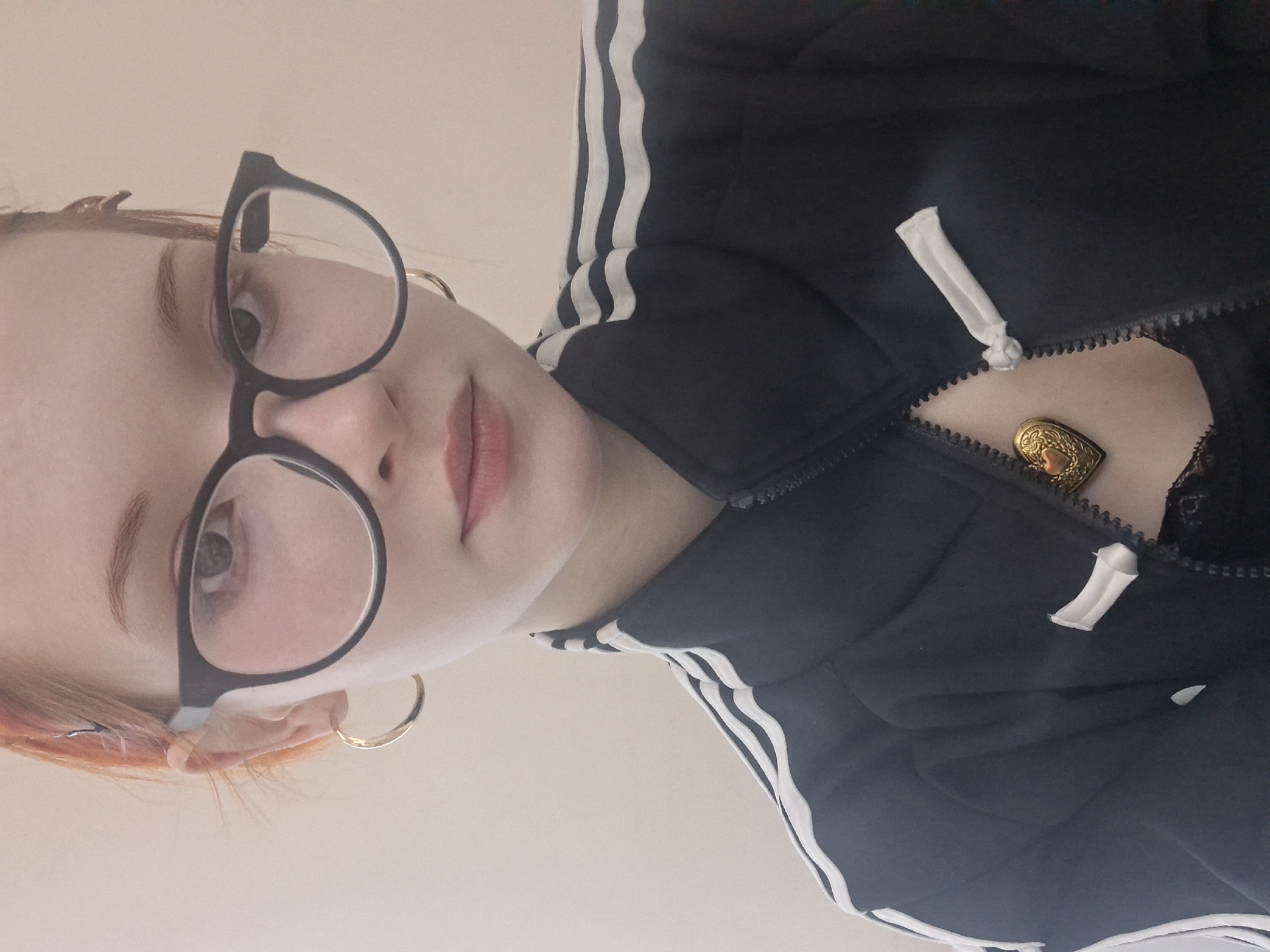 A selfie taken from a low angle features a person with fair skin, light reddish-blonde hair, and light-colored eyes. They are wearing round, black-framed glasses and gold hoop earrings. The person has their lips slightly parted. They are wearing a black zip-up jacket with three white stripes on the sleeves. The jacket is partially unzipped, revealing a glimpse of black lace underneath and a small, gold heart-shaped object visible inside the jacket. The background appears to be a plain, light-colored wall. There is nothing in the image to indicate the location.