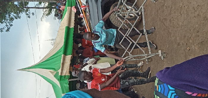 The image shows a group of people standing in front of a large tent and a black vehicle. The coordinates 0.562, 34.805 place the scene in Kivaywa, Kenya, which is located in the Bungoma County. A man in a blue shirt and black pants is pushing a foldable metal cart towards the vehicle. Another man in a red shirt and black pants is standing on the left side of the cart. They are both wearing black shoes. In the background, there are other people, some of whom are standing and some of whom are seated. The vehicle's tire is visible in the background. The ground is dirt. The sky is overcast. The image appears to have been taken during a gathering or event, likely in a rural or semi-rural area. The presence of the foldable metal cart and the vehicle suggests that this could be a gathering for a social or ceremonial event.