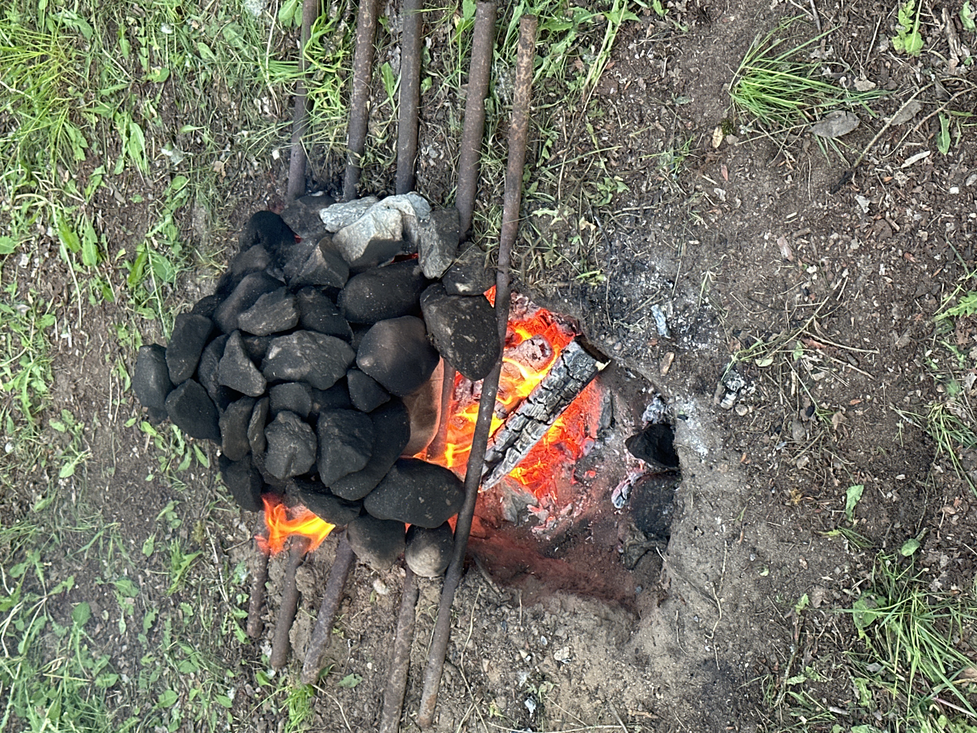 The image depicts an outdoor fire pit in Папярнянскі сельскі Савет, Belarus. The pit is dug into the earth, with charred remains and ash visible. A fire is burning with visible flames and glowing embers. A collection of black rocks, with a few grey ones mixed in, is piled on top of a metal grate or series of bars over the fire. Patches of green grass and bare earth surround the fire pit. There are no people visible.
