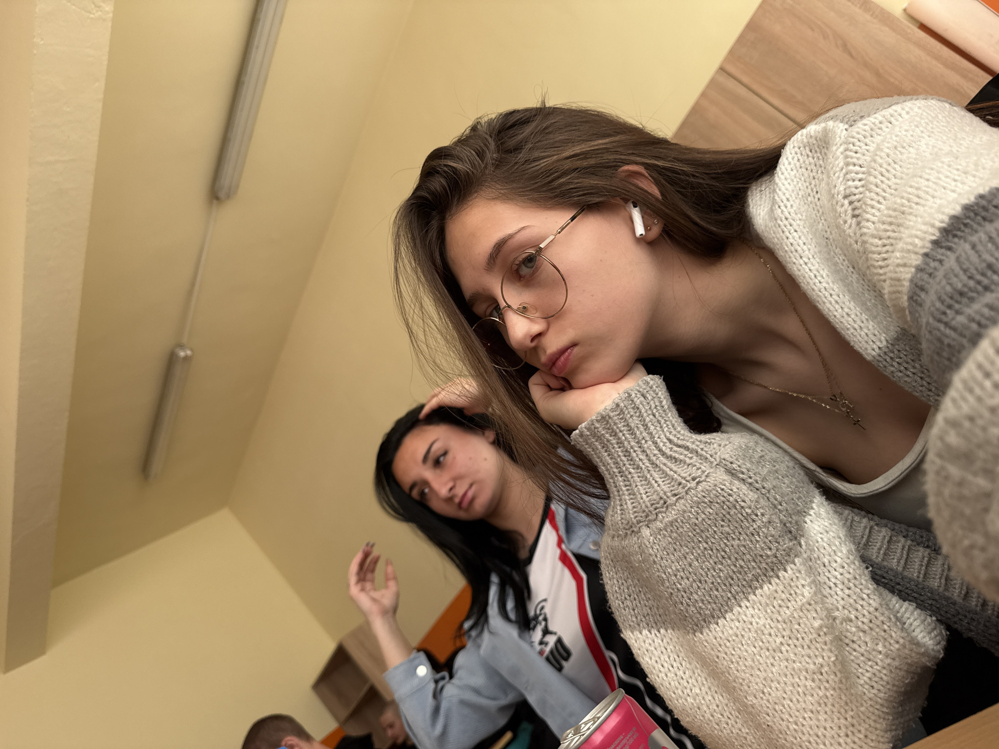 An eye-level shot captures a casual indoor scene with three individuals, likely in a classroom or study area in Sofia, Bulgaria. The main subject, a young woman with long brown hair, round gold-rimmed glasses, and AirPods in her right ear, occupies the foreground. She wears a cream and grey striped knitted cardigan over a light top, revealing a delicate gold necklace. Her chin rests on her hand, conveying a pensive or bored expression as she looks slightly downwards.
In the mid-ground, a second young woman with long dark hair is partially visible and out of focus. She wears a light blue denim jacket over a top with white, red, and black details, and one hand is raised near her head. The back of a third person's head, wearing blue headphones, is faintly seen in the lower-left corner.
The setting features light yellow/beige walls and a ceiling with two fluorescent tube lights. A wooden desk or table surface is visible on the right, and a shelving unit can be seen in the blurred background. A pink can, possibly a beverage, with partially visible text, rests in the lower-center foreground. The overall atmosphere suggests a moment of quiet or pause during an activity.