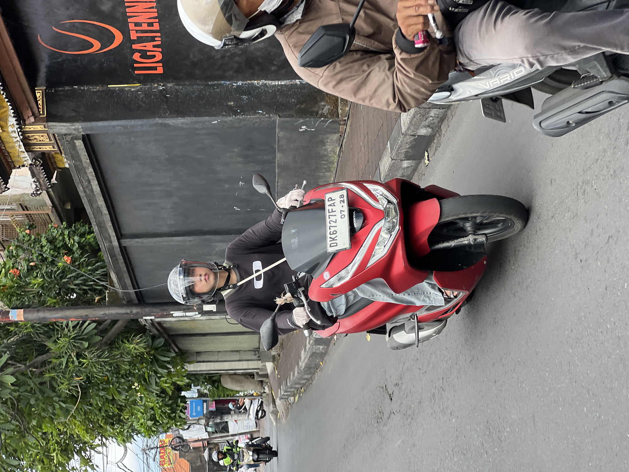 In North Kuta, Indonesia, on a paved street, a young person wearing a helmet and gloves rides a red scooter. The scooter's license plate reads "DK6727FAP" and "07-28". The rider is wearing a black long-sleeved shirt and light blue jeans. Behind this scooter, another person on a scooter, wearing a brown jacket and a face mask, is partially visible. A black wall with an orange logo and text reading "LIGA.TENNI" is seen on the right side of the frame. Lush green trees line the street. The sky appears overcast.