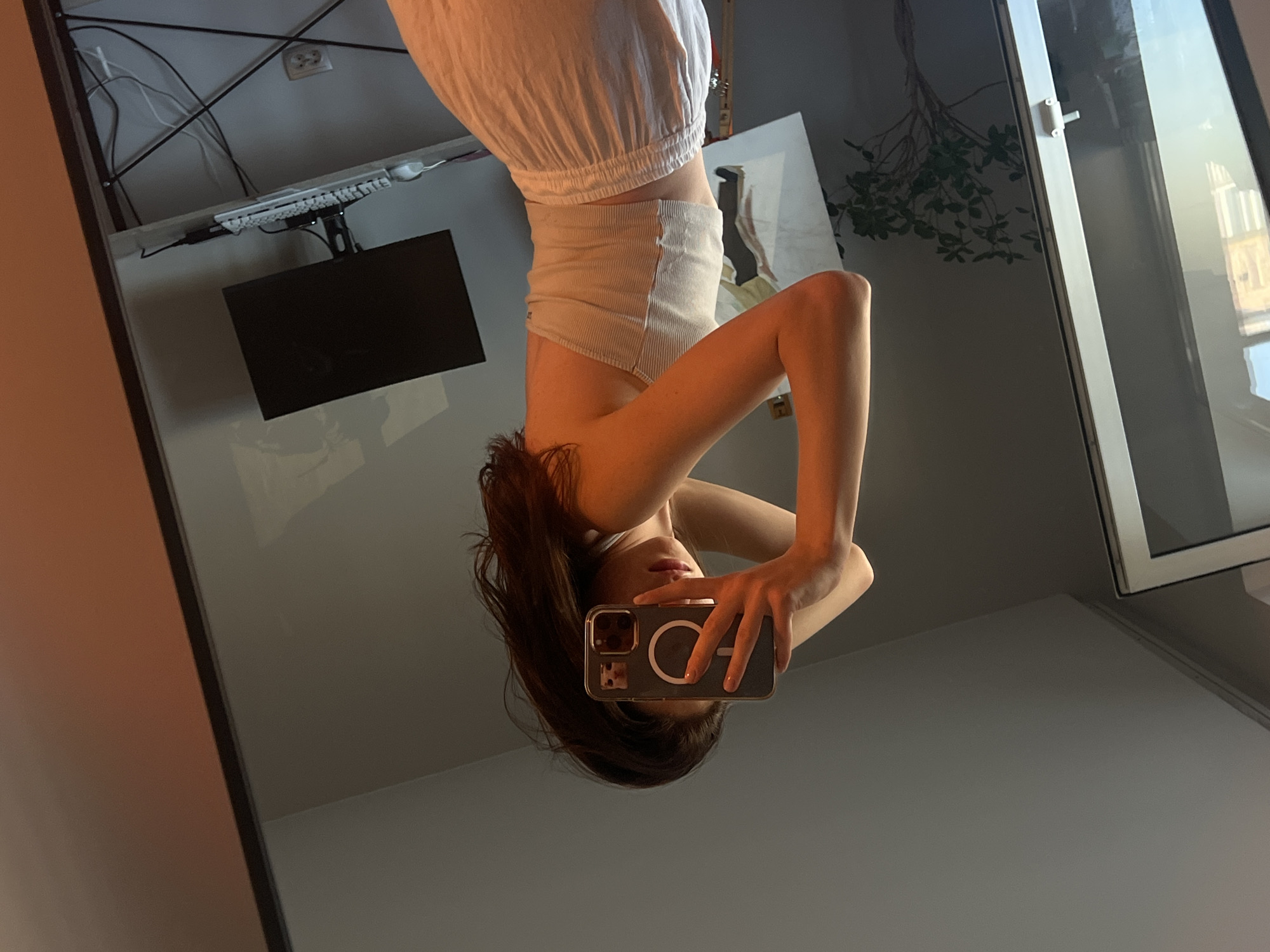 The image is a wide-angle, upside-down, mirror selfie taken indoors. The primary subject is a young woman, who appears to be in her late teens or early twenties, posing for the camera. She is wearing a white, ribbed tank top and white shorts or a skirt. Her dark hair is falling forward, obscuring part of her face. She is holding a smartphone in her right hand, angled to capture the reflection. The phone case is dark with a prominent circular magnetic accessory.
The setting appears to be a home office or study area. In the background, reflected in the mirror, there is a desk with a computer monitor mounted on an arm. A keyboard and various cables are visible on the desk. The walls are a neutral gray, and a large window is visible on the right side of the frame, suggesting it might be daytime. A hanging plant with green leaves is also reflected in the window, adding a touch of nature to the indoor scene. The lighting suggests it might be late afternoon or early evening, with a warm, soft glow, possibly from the sun. The overall mood is casual and relaxed, possibly taken during a moment of leisure at home in Ternopil, Ukraine. There is no visible text in the image.