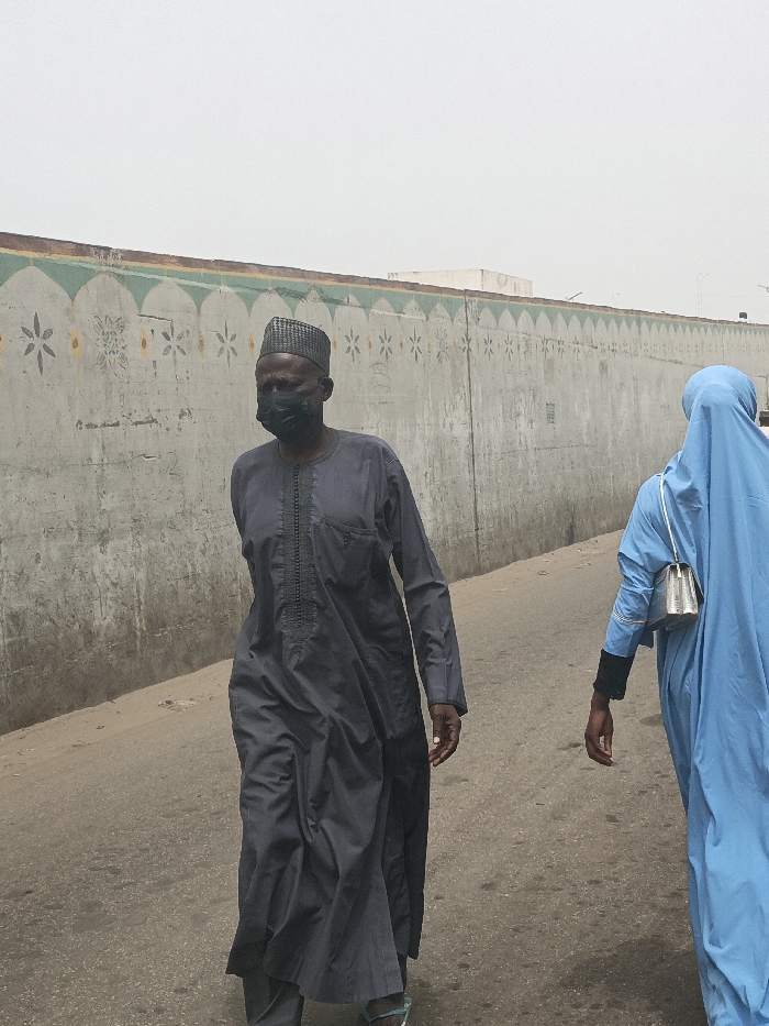 In Fagge C, Nigeria, a daytime outdoor scene depicts two individuals walking on a paved, grayish-brown surface. An adult male, positioned centrally, walks leftward, wearing a dark gray long-sleeved tunic with vertical embroidery, a dark patterned cap, and a black face mask. His hands are at his sides, and light blue sandals are visible. Behind and to the right of the male, an adult female walks in the same direction, primarily seen from her back and right side. She wears a light blue abaya or hijab covering her head and body, with a black long-sleeved garment visible on her left arm. A silver-colored handbag with a white strap is slung over her right shoulder. In the background, a long, weathered gray concrete wall features a decorative upper frieze consisting of green arched patterns alternating with yellow floral and star-like motifs. A light-colored, flat-roofed structure is partially visible above the wall. The sky is uniformly overcast and light gray.