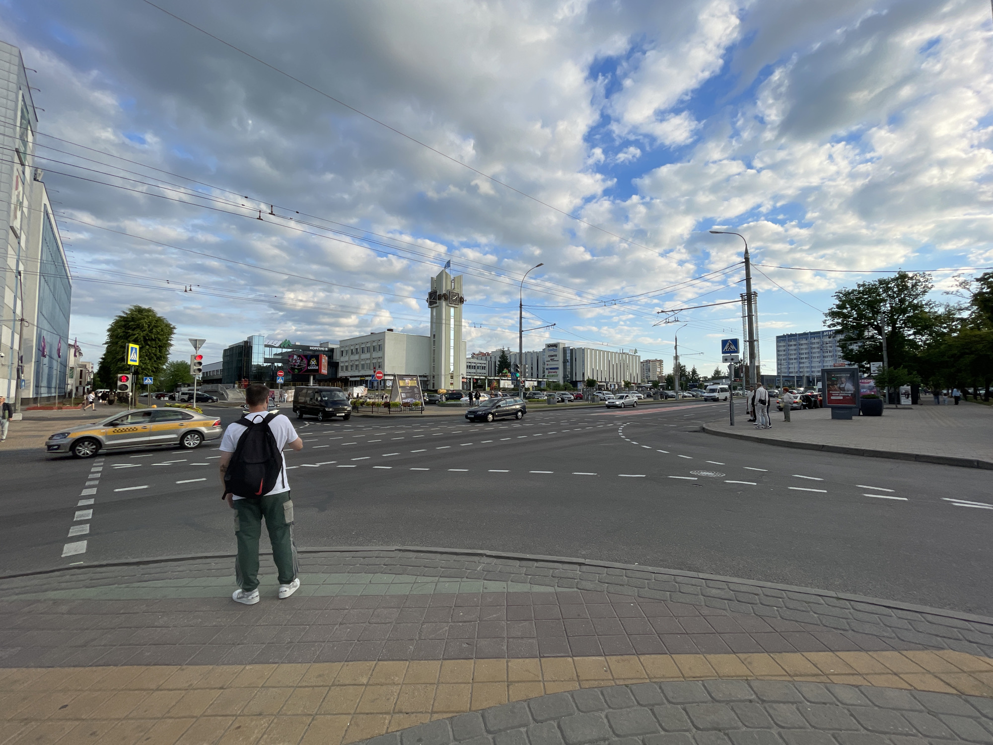 This image depicts a wide-angle street scene at a busy urban intersection in Brest, Belarus, specifically at the intersection of Gogolya Street and Sovetskaya Street, identifiable by the distinctive clock tower. The time of day appears to be late afternoon or early evening, under a partially cloudy sky with patches of blue.
In the immediate foreground, a tiled sidewalk in shades of grey and yellow transitions to a curb marked with a green stripe, indicating an accessible ramp for a pedestrian crossing. A male figure is positioned centrally, viewed from behind, standing at the edge of this curb. He wears a white t-shirt, dark green cargo pants, and white athletic shoes, with a black backpack (featuring red accents) on his back. His right arm is slightly raised.
Directly in front of the pedestrian, the asphalt roadway is marked with white dashed lines forming a crosswalk. Further into the intersection, additional white lines define traffic lanes and stop points. A silver Volkswagen sedan, marked as a taxi, is turning left across the crosswalk from the left side of the frame. A red traffic light is visible above this taxi. Other vehicles, including a dark minivan and a dark hatchback, are present within the intersection and on the surrounding streets.
To the left, a modern building with glass and light-colored paneling stands, displaying an advertisement on its lower section. In the midground, towards the center-left, a prominent, tall, slender clock tower with multiple clock faces and a blue flag flying from its apex is visible. This is the landmark clock tower on Gogolya Street. Adjacent to and beyond the clock tower, various commercial and residential buildings line the streets, some featuring advertisements such as "Ozon." Streetlights and a network of overhead wires (likely for trolleybuses or power) traverse the scene. Green foliage from trees is scattered throughout, particularly on the left and right sides of the image.
In the background, a dense urban environment continues with more multi-story buildings, including a tall, light blue-grey building on the far right. The sky is marked by large, fluffy white cumulus clouds, interspersed with clear blue sky, casting a soft, even light over the scene. Several other pedestrians are visible in the distance on sidewalks.