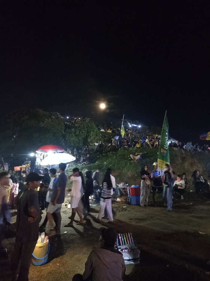 This nighttime photograph captures a large, bustling outdoor gathering on and around a grassy hillside in Kecamatan Pujut, Indonesia. The dominant feature of the dark sky is a prominent, bright full moon, illuminating the scene.
In the foreground, numerous people are standing, walking, and engaging with vendors. To the left, a food or merchandise stall with a red and white striped umbrella is brightly lit, attracting a small crowd. Another individual is seated in the lower right foreground, tending to a basket filled with what appear to be sparklers or small fireworks.
The midground shows more individuals interspersed with two large green flags. One flag is clearly visible, displaying the text "Bank" and partially "Bri," indicating it's likely a Bank BRI advertisement. The flags are positioned on a gentle slope leading up to the main gathering area.
The hillside in the background is densely populated with a multitude of people, many of whom are seated or standing, observing the surroundings. Small tents or shelters are visible among the crowd on the slope, along with scattered lights. The atmosphere appears lively and social, suggesting a public event, festival, or a communal viewing of the moon. The clear night sky and the visible moon imply favorable weather conditions.