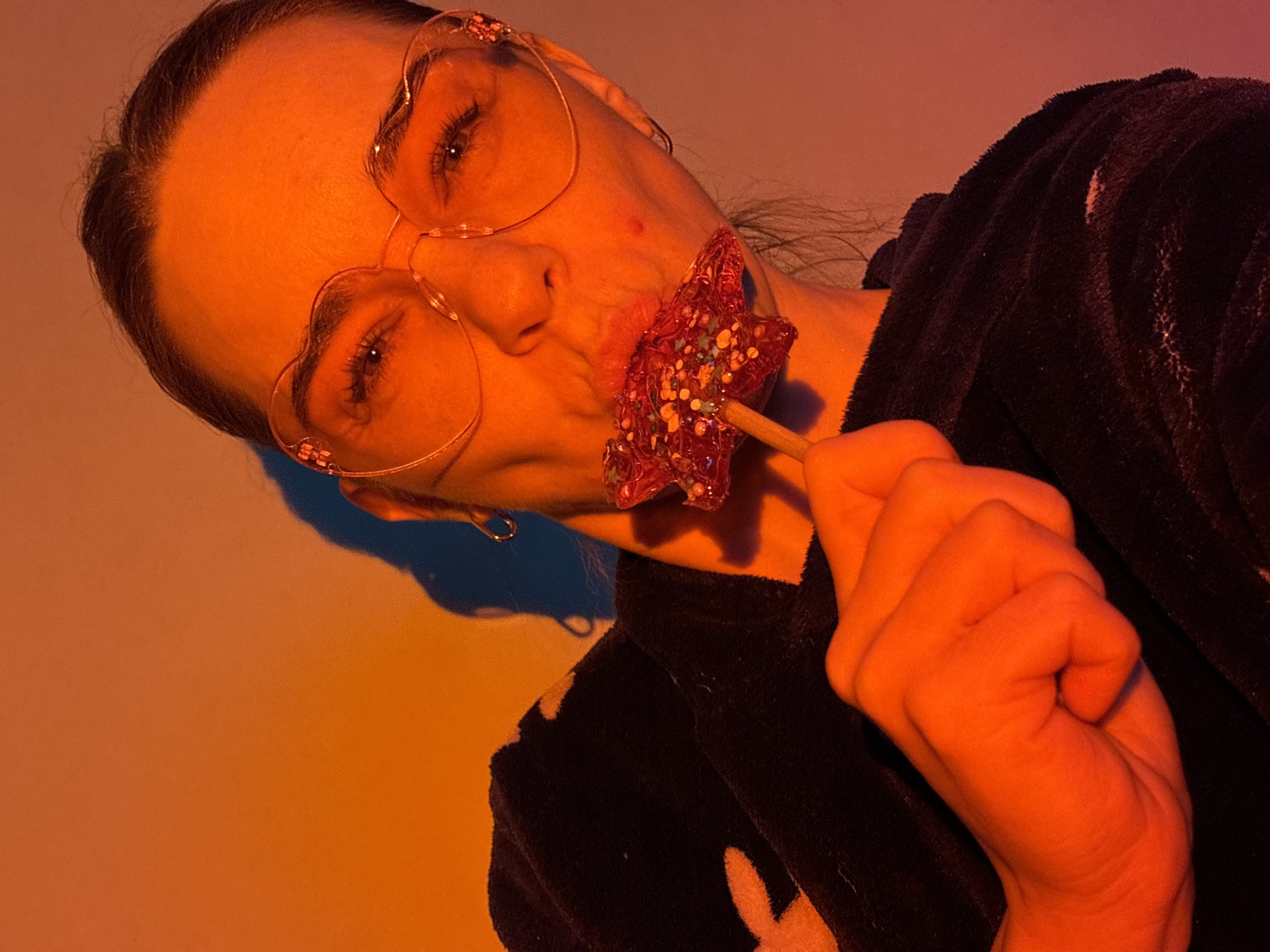A woman with fair skin and pulled-back hair is angled towards the viewer, wearing heart-shaped glasses and a dark, patterned robe. She is holding a star-shaped lollipop with sprinkles to her mouth. The scene is lit with an orange hue, possibly from artificial lighting. The location is given as Unknown, Russia.