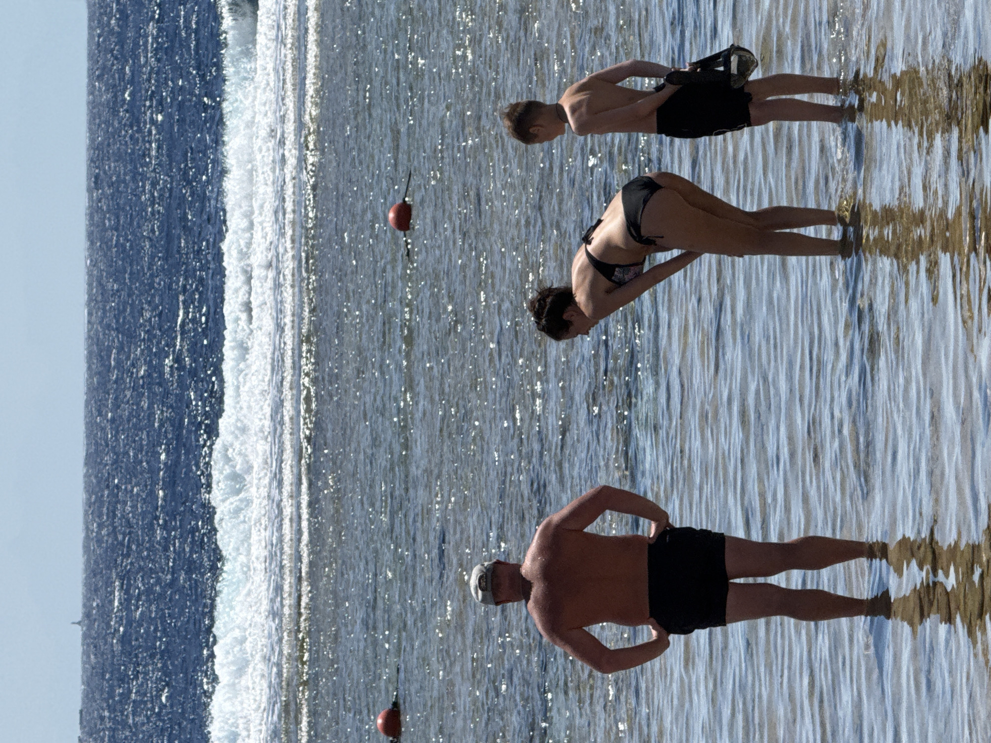The image depicts three individuals near the shoreline of a body of water, possibly the Red Sea in Sharm El Sheikh, Egypt. The scene is bathed in bright sunlight, suggesting it is daytime, likely mid-morning or afternoon, with clear weather.
In the foreground, a shirtless man wearing black shorts and a cap stands with his hands on his hips, facing away from the camera. His reflection is visible in the rippling water. Behind him, a woman in a black bikini is captured in a dynamic pose, appearing to be in mid-motion, possibly performing an exercise or dive. She is facing away from the camera as well. Further back and to the left, a younger person, likely a boy, is also facing away, wearing black shorts and holding a dark object.
The water is a prominent element, with a line of white surf breaking close to the shore, indicating a shallow area transitioning to deeper water. The surface of the water is highly reflective, creating a shimmering effect due to the sunlight. A red buoy with a black rope is visible floating in the water between the two individuals in the background. The overall atmosphere appears relaxed and leisurely, typical of a beach or coastal resort setting. No visible text is present in the image.