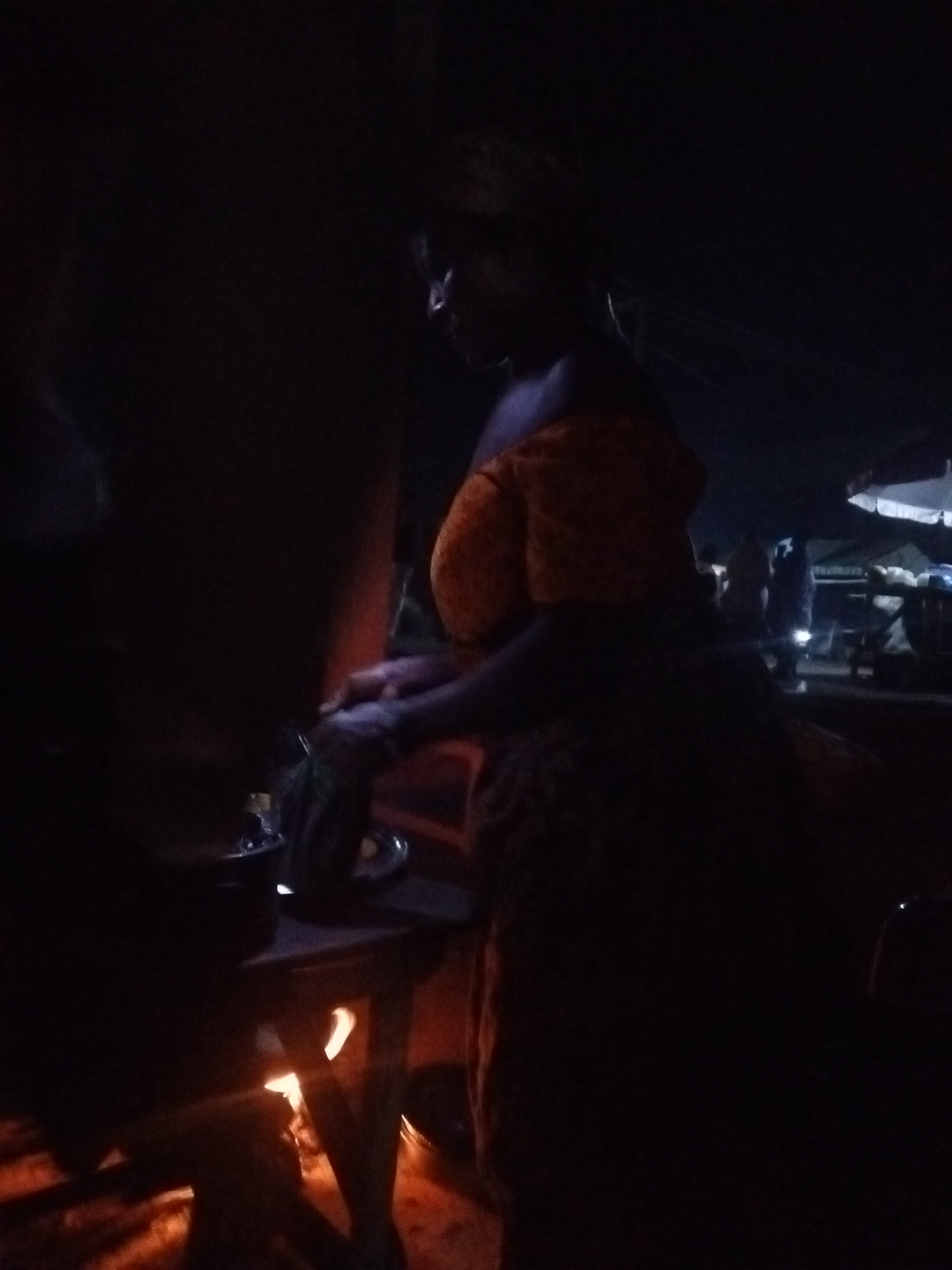 The image depicts a dimly lit outdoor scene at night, likely in a market or roadside setting. A woman, partially illuminated and wearing an orange off-the-shoulder top and a patterned skirt, is standing at a low wooden table. Her hands are engaged with something on the table, possibly preparing food or an item for sale. A fire is visible beneath the table, casting a warm glow and creating a sense of ambient light. In the background, other figures and structures are faintly visible, suggesting the presence of other stalls or people. The overall atmosphere is one of activity in a low-light environment.