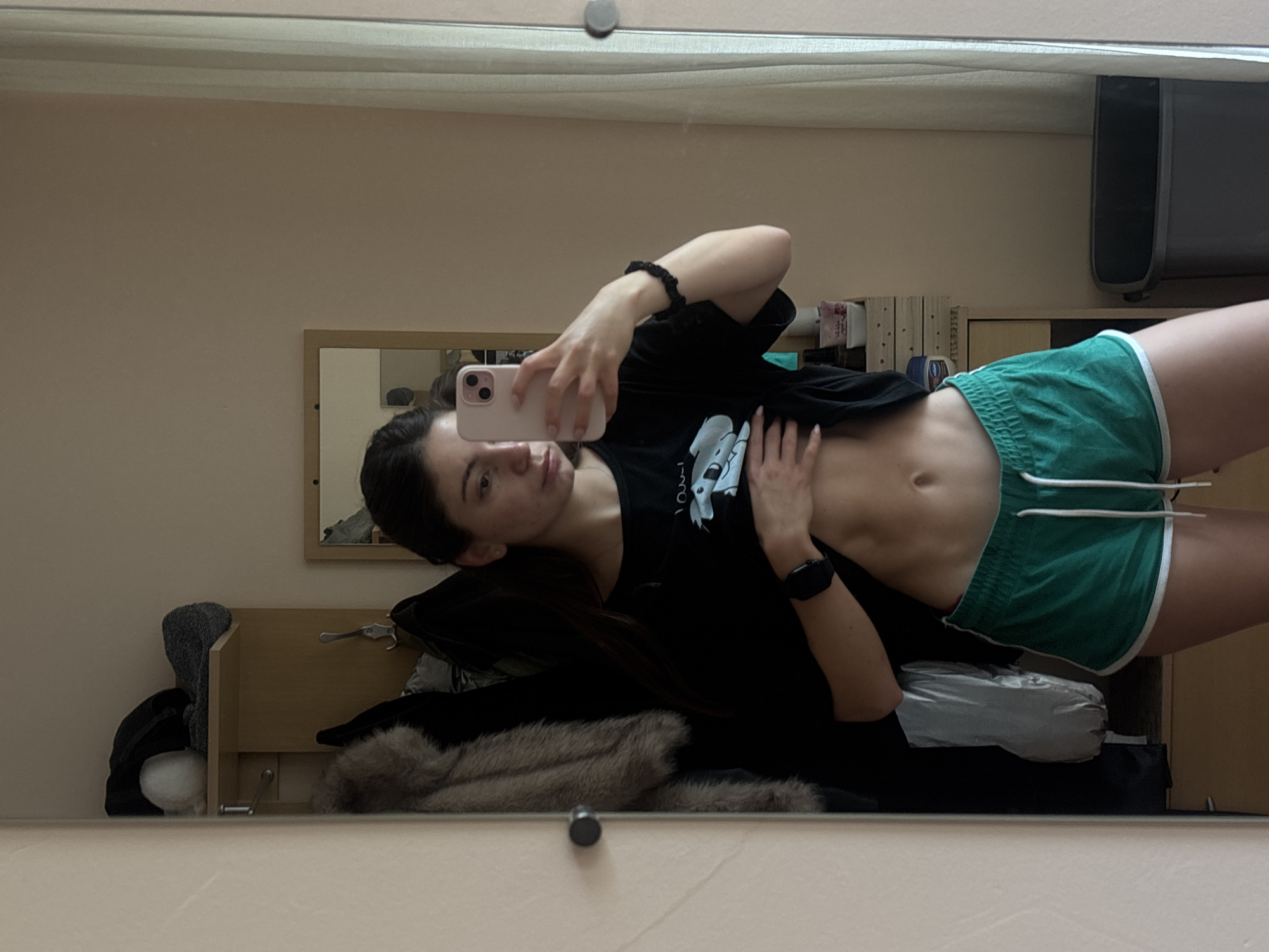 A young woman with long dark hair is taking a mirror selfie in an apartment. She is wearing a black t-shirt and green athletic shorts. Her t-shirt is slightly lifted to reveal her toned abdomen. She is holding a pink smartphone in her right hand to take the photo. A black smartwatch is visible on her left wrist. In the background, a closet door is open, revealing a fur coat and other clothing items. A small mirror is hanging on the closet door, and a shelf above it holds various items. A trash can is visible to the right of the woman. The walls are painted a pale pink color. The overall impression is one of a casual, personal moment captured within a home environment. There is no indication of the location being Varna, Bulgaria.