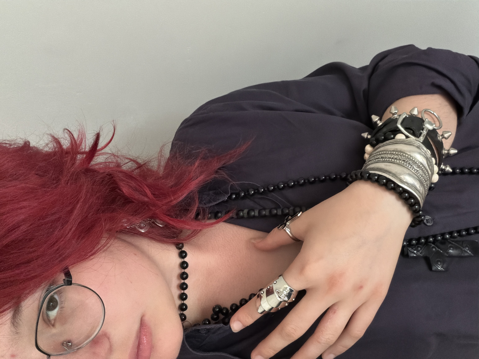 The image shows a close-up of a person with bright red hair and glasses, wearing a dark shirt and multiple pieces of jewelry. The person is wearing a choker necklace made of black beads, a longer strand of black beads, a large silver ring on their index finger, and several stacked bracelets on their wrist. The bracelets include a silver cuff with intricate carvings, a black leather band with silver spikes, and other beaded bracelets. The person's left hand is resting on their chest, and their right hand is partially visible. The background is a plain, light-colored wall. There is no visible indication of the location being in Chelyabinsk, Russia, or any other specific city.