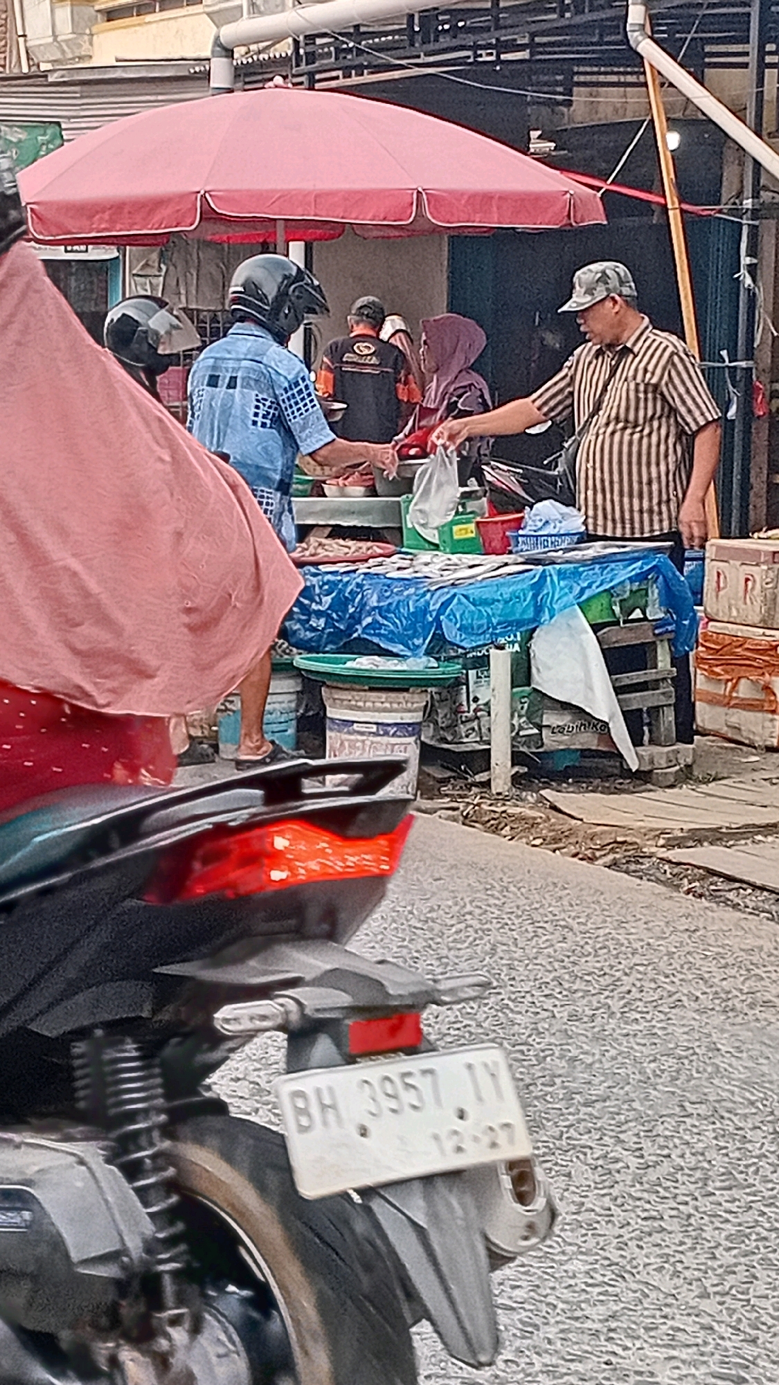A medium shot shows a street market scene in Jambi, Indonesia, with people interacting at a stall. In the foreground, the rear of a motorcycle with a license plate reading "BH 3957 1Y 12-27" is visible. The market stall, covered by a red umbrella, displays fish on a table draped with a blue tarp. A person wearing a blue checkered shirt and a black helmet is handing something to another person wearing a striped shirt and a camouflage cap. Other individuals are present in the background, some obscured. The environment appears to be an outdoor marketplace with stalls and a road.