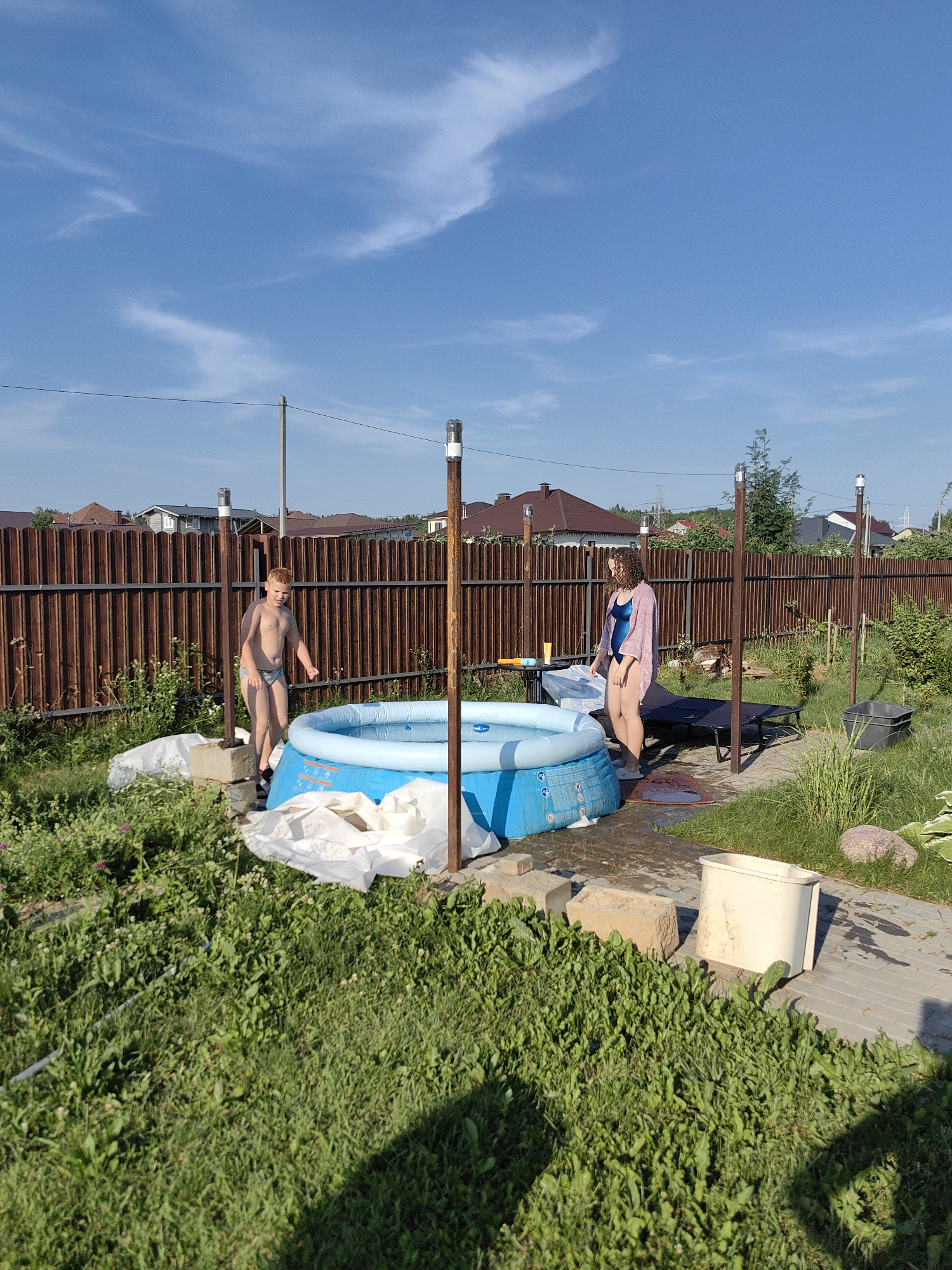 A young boy and a woman are in a backyard with an inflatable pool. The boy, wearing swimming trunks, stands near the pool, looking towards the viewer with a slight smile. The woman, wearing a blue swimsuit and a light pink cover-up, stands next to a lounge chair, looking down at the pool. The inflatable pool is blue and partially filled with water. In the background, a wooden fence encloses the yard, with houses and trees visible beyond. The scene appears to be set on a sunny day, with a blue sky and scattered white clouds, suggesting it is daytime. There are several objects in the yard, including a lounge chair, a bucket, bricks, and some white tarps. The overall mood seems relaxed and summery. The location is identified as Fanipal, Belarus. There is no visible text in the image.