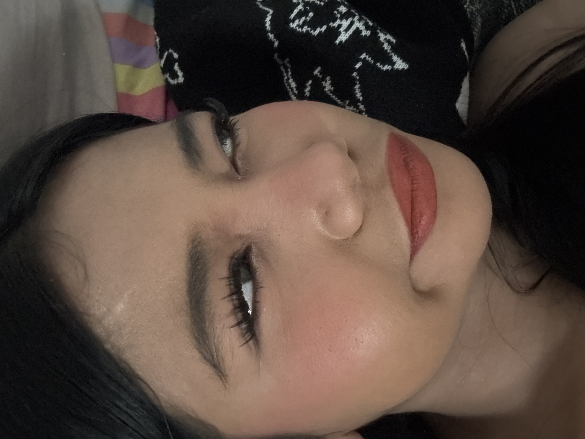 A close-up, eye-level shot focuses on the right side of a young woman's face. Her head is tilted, and her gaze is directed upwards and to her right. She has dark hair that falls to her forehead and right shoulder. Her skin tone is medium, and she is wearing makeup. Her eyebrows are dark and well-defined. Her right eye is open, with long, dark eyelashes. The iris appears to be dark brown. Her left eye is mostly obscured by her head tilt, but a sliver of her iris is visible, also appearing dark. She has prominent cheekbones, with a dusting of blush on her right cheek. Her nose is short and slightly upturned. Her lips are full and painted with a matte, coral-red lipstick. Her chin is slightly turned, creating a subtle dimple. The background is out of focus, with a striped pattern of pink, yellow, purple, and white visible on the left, and a black garment with a white embroidered pattern on the right. The lighting suggests an indoor setting. No specific location in North Jakarta, Indonesia can be determined from the image.