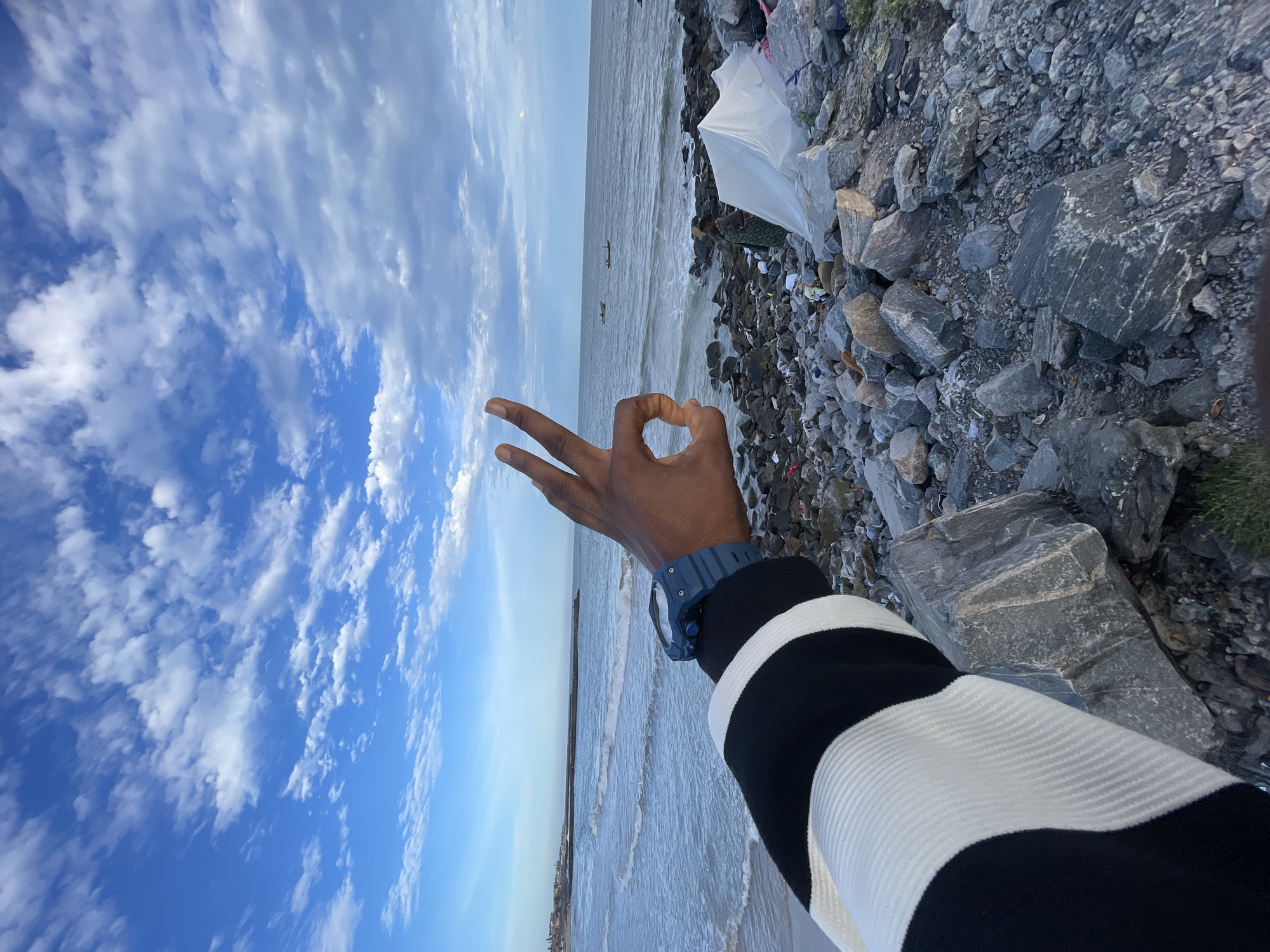 The scene is an outdoor shot, likely taken from a beach, featuring a person's hand making a gesture in the foreground. In the background is the beach and sea and along the coast is trash. The sky above is filled with white clouds against a blue sky. The location is Alaguntan, Nigeria.