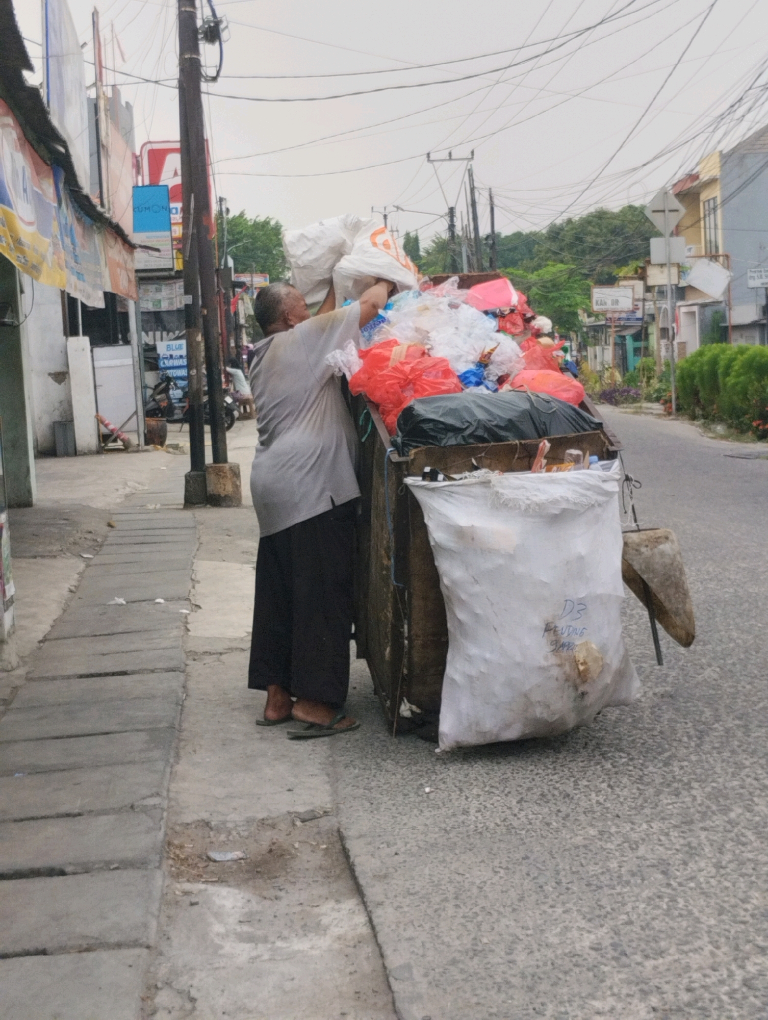In Bekasi, Indonesia, a person wearing a gray t-shirt and dark pants is tending to a large cart overflowing with plastic bags of collected waste. The cart, seemingly homemade and constructed from wood, is positioned on the side of a paved street. The person is actively adding more items to the already full cart, with visible red and white plastic bags dominating the load. A large white sack is tied to the front of the cart, bearing handwritten blue markings. The scene unfolds in a street environment with buildings and utility poles lining the road, suggesting an urban or suburban setting. The pedestrian walkway, made of concrete slabs, runs alongside the street. The sky appears overcast.