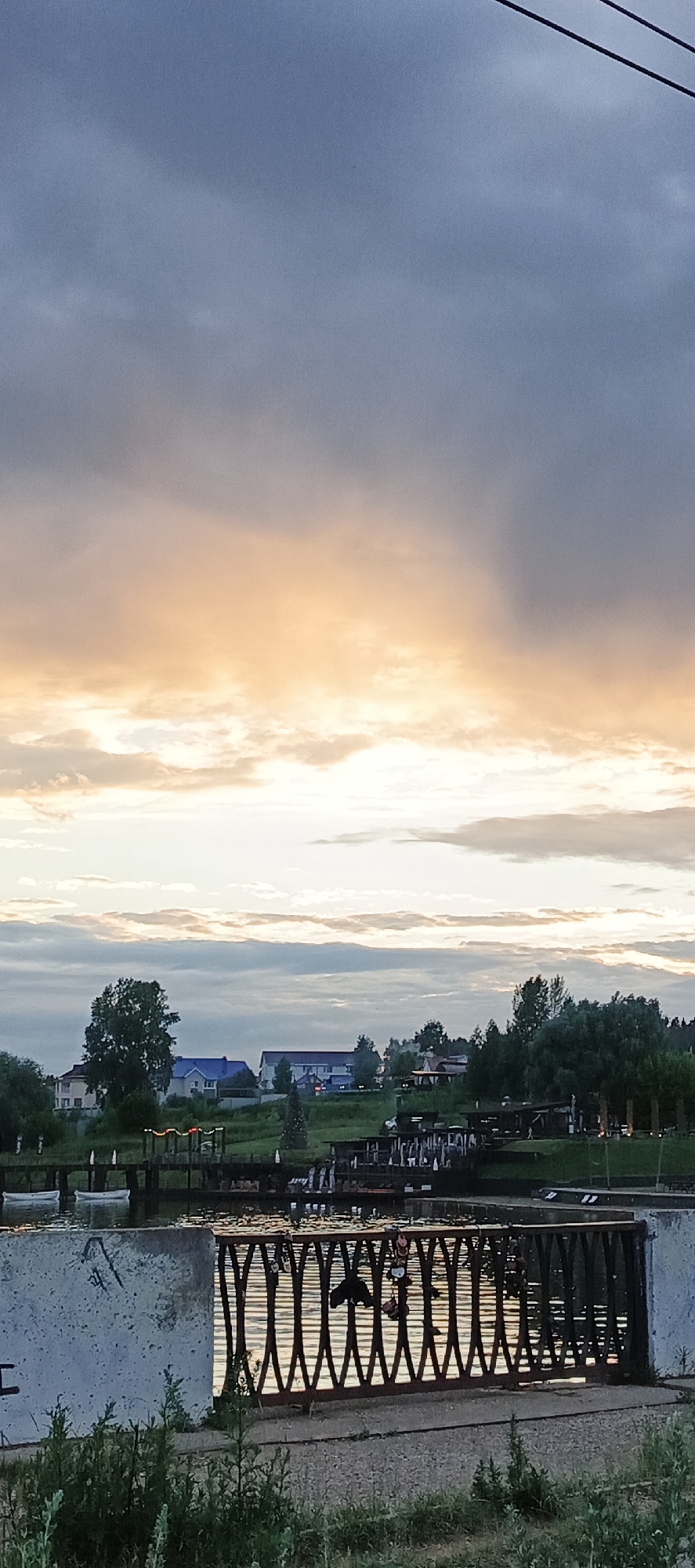 The image depicts a waterfront scene at dusk in Votkinsk, Russia. The sky is filled with dramatic clouds, transitioning from a deep blue at the top to warm oranges and yellows near the horizon, suggesting either sunrise or sunset. In the middle ground, a line of trees and houses with blue roofs are visible on a grassy embankment. A body of water separates the viewer from the embankment, with several small boats moored along the edge. In the foreground, a concrete barrier is visible on the left, and a dark metal railing with a decorative, wavy pattern runs across the bottom of the image. This railing has several love locks attached to it, a common practice in many locations to symbolize commitment. The water reflects the colors of the sky and the railing, creating a shimmering effect. The overall atmosphere is serene and tranquil, characteristic of a quiet evening by the water.