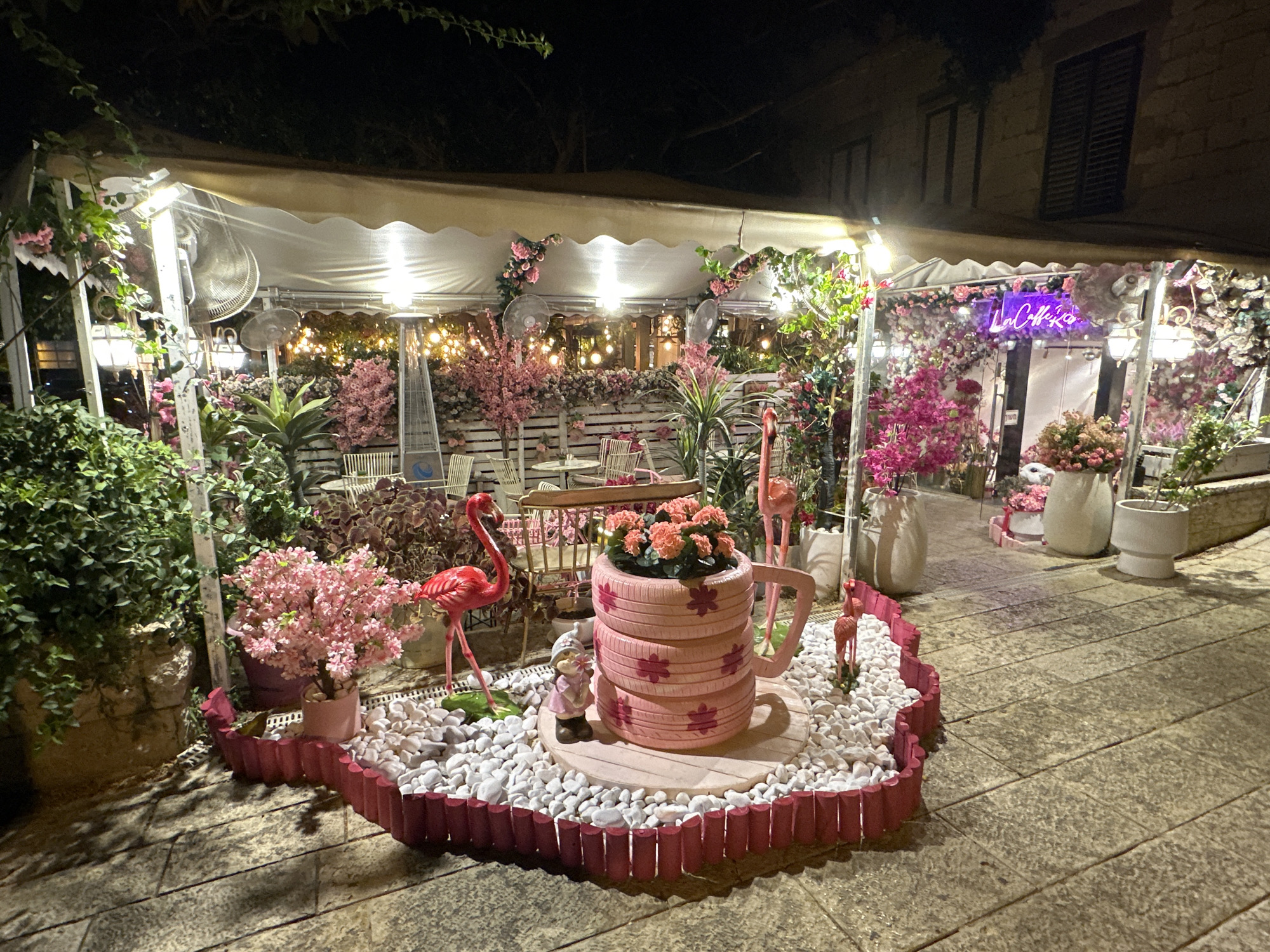 The image depicts a charming outdoor seating area at night, likely a cafe or restaurant, adorned with an abundance of pink flowers and vibrant decorations. The scene is set on a paved walkway, with a building visible in the background. Strings of warm-toned lights are draped across a white canopy, illuminating the space and creating a festive ambiance.

Prominently featured is a large, pink, tiered planter shaped like a mug, filled with coral-colored flowers. Beside it, a collection of pink flamingo lawn ornaments adds a playful touch, with one larger flamingo standing guard near the mug planter and smaller ones scattered around. A small gnome figurine is also present near the planter. Lush green foliage spills from various pots and hangs from the canopy, intermingling with the artificial pink flowers that festoon the area. White chairs and tables are visible under the canopy, suggesting a seating area. In the background, a neon sign with purple lettering reads "La Collina". A patio heater stands to the left, hinting at cooler evening temperatures. The overall impression is one of whimsical delight and an inviting atmosphere for patrons. The location context is Haifa, Israel.