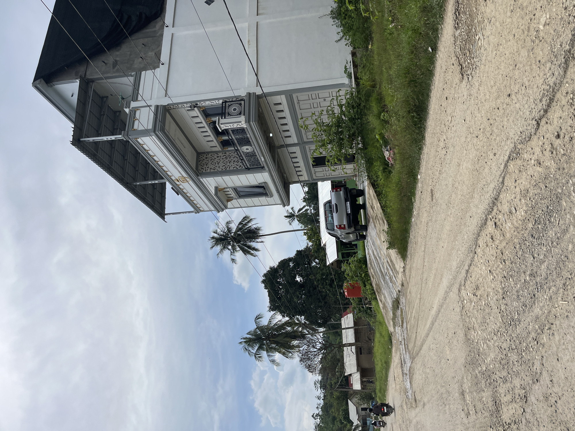 The image depicts a cloudy day in a rural area of Perupuk, Indonesia. In the foreground, a dirt road with visible tire tracks and potholes stretches into the distance. To the right, a modern, multi-story white house with grey trim and balconies stands prominently. In front of the house, a silver pickup truck is parked on an incline leading to the road.
Further down the road, several smaller, simpler houses with corrugated metal roofs are visible, some appearing to be made of wood. Two individuals on motorcycles are riding on the dirt road, heading away from the viewer. In the background, lush green vegetation, including several palm trees and dense foliage, covers the landscape. A large red water tank is situated near one of the smaller houses. The overall impression is one of a developing rural community.