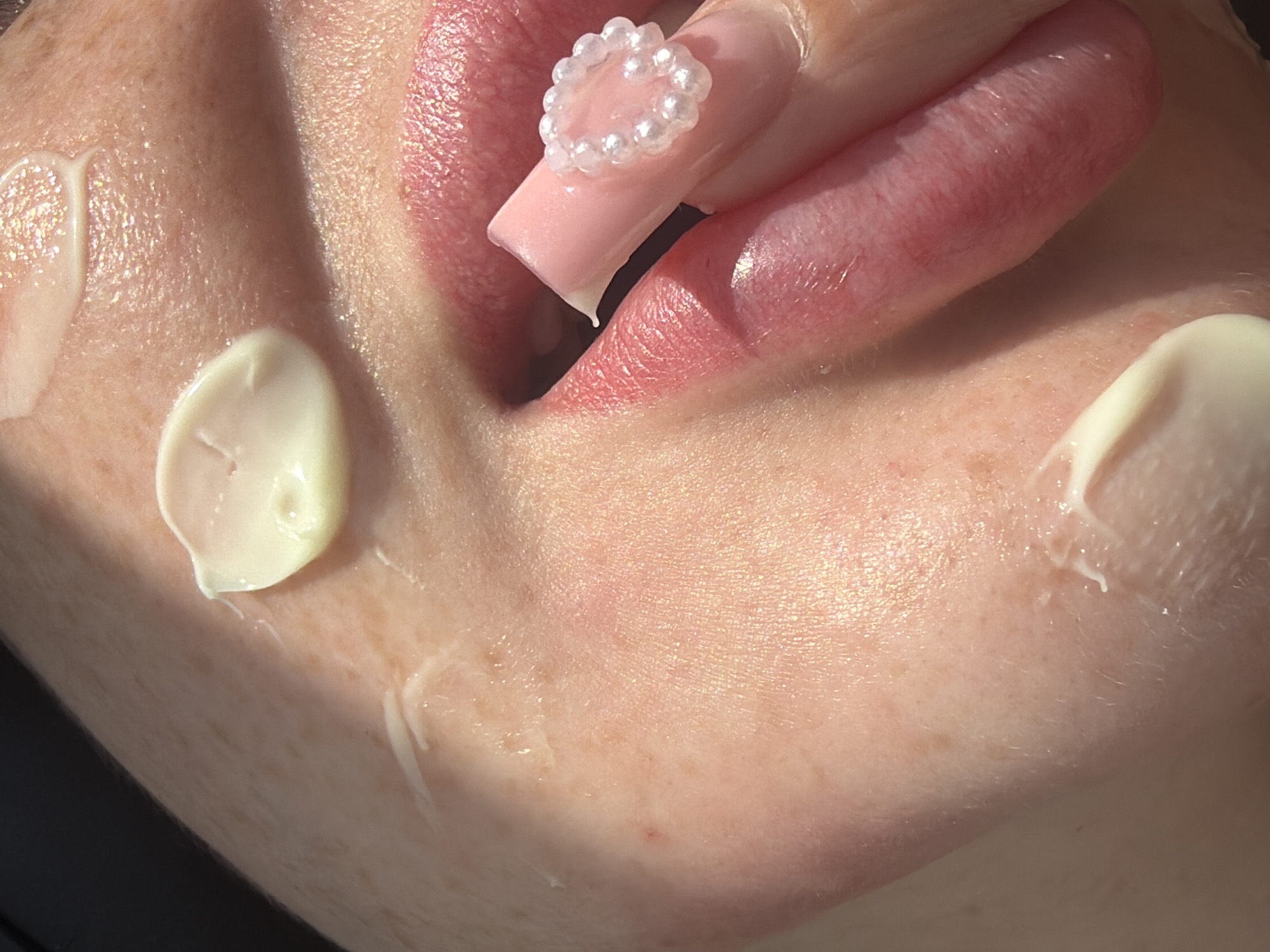 The close-up image shows a person's cheek and lips with a cream or lotion applied to the skin. There are several dabs of the product, with one dab prominently placed on the cheek. The person's lips are slightly parted, revealing a pinkish hue. A fingernail adorned with small white beads is visible near the lips, and a small amount of the cream is on the nail. The skin has freckles and a slightly shimmery appearance, possibly due to the product. The background is dark, creating a strong contrast with the illuminated skin. The image does not provide any information about the location in Saint Petersburg, Russia.
