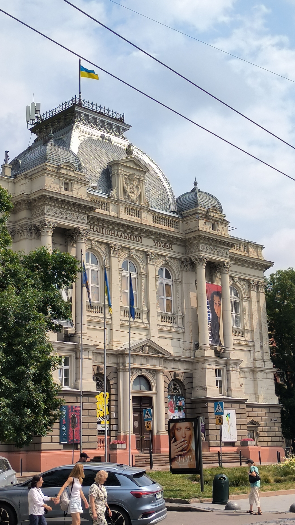 The image depicts the National Museum in Lviv, Ukraine, under a partly cloudy sky. The grand facade of the museum is a prominent feature, characterized by classical architecture with columns, arched windows, and decorative elements. A Ukrainian flag flies from a flagpole atop the building's roof. Several other flags, including what appears to be the European Union flag, are also displayed near the entrance.
In front of the museum, a sidewalk and street are visible, with trees lining the building. Several posters and banners are displayed on the museum's exterior, advertising exhibitions or events. One banner features portraits of people, and another has text in Ukrainian. On the sidewalk, a gray car is parked, and several people are walking. One group of three women is walking together, while a single woman stands on the sidewalk near a trash can. Another person is visible in the background, walking away from the camera. The scene appears to be a public space in Lviv, likely a busy day with pedestrian and vehicular activity.