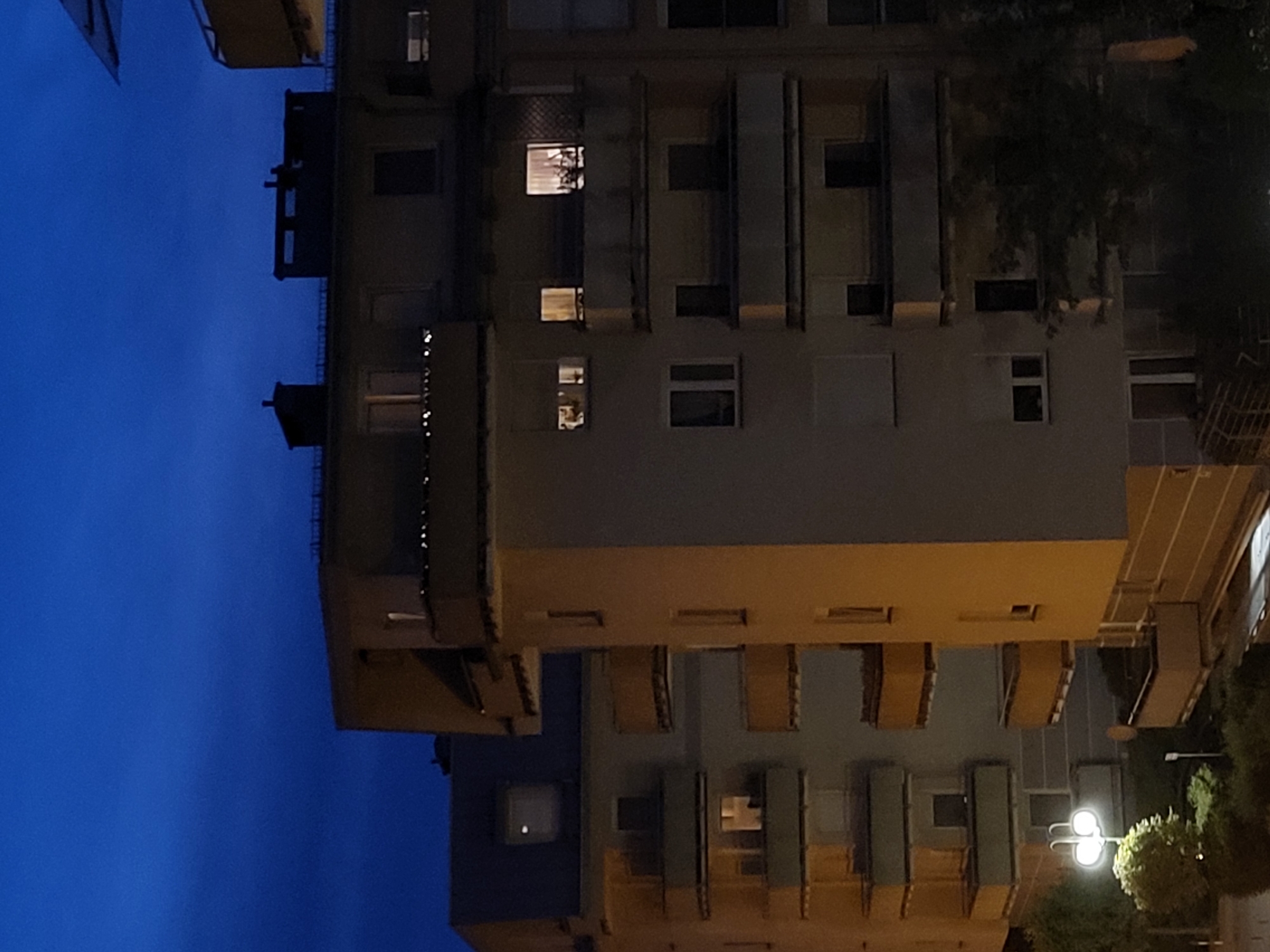 A nighttime, exterior shot shows apartment buildings against a dark blue sky. The buildings are illuminated by interior lights and a streetlamp. The buildings have multiple stories and balconies with some railings and yellow accents. Some windows are lit, suggesting occupancy. A streetlamp with two globes is visible in the foreground, illuminating some trees and bushes below. The overall scene suggests an urban residential area at night.