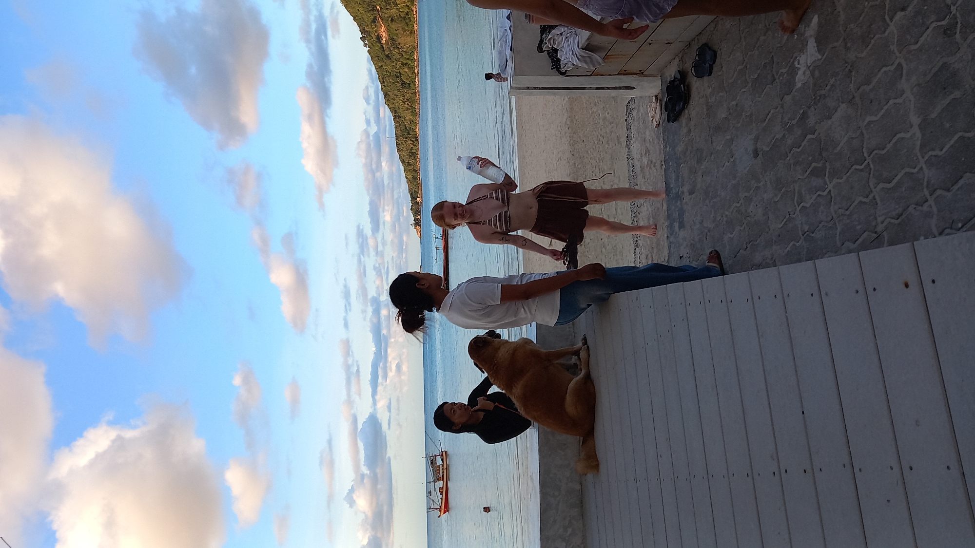 In ตำบลเกาะพะงัน, Thailand, on a beach, two women are standing on the sand near the water's edge. One woman, with red hair, is wearing a striped bikini top and brown shorts, holding a clear plastic bottle. The other woman, with dark hair pulled back, is wearing a white polo shirt and blue jeans, facing the first woman. A large, light brown dog is sitting on a wooden deck to the left, with another woman in a black long-sleeved shirt behind it, looking towards the two women on the sand. In the background, the ocean is calm with two boats visible on the water. The sky is partly cloudy with a mix of white and gray clouds against a blue sky. A tree-covered hillside is visible on the right side of the frame. The foreground consists of a wooden deck and a paved area with several pairs of flip-flops scattered on the ground.