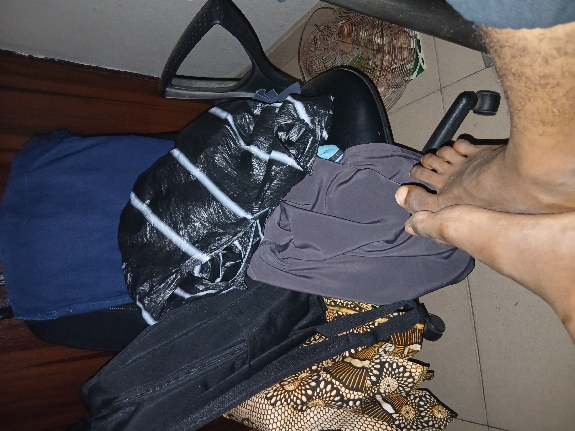 A view from above of a swivel chair covered with clothes and a person's bare feet resting on top of the clothes. The chair is black and has a back with a U-shaped opening. On the chair are a blue shirt, a black plastic bag with white stripes, a gray cloth, a black jacket with a zipper, and a bag with a pattern of white and black flowers on a brown background. The floor is tiled. An electric fan is visible in the background. The location is Unknown, Unknown.