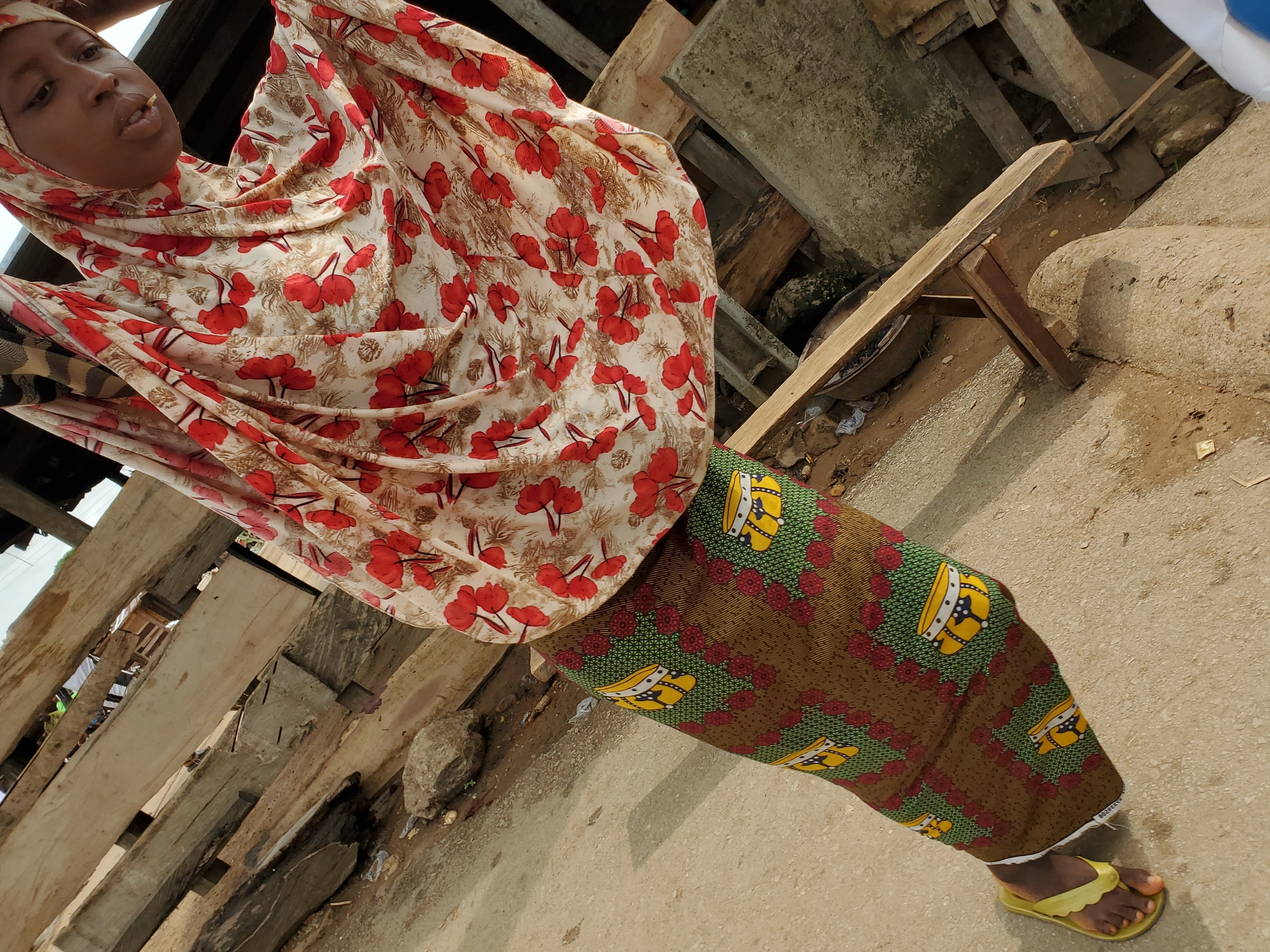 A person stands on a concrete surface in what appears to be an outdoor environment in Mararaba, Nigeria. The individual is wearing a head covering featuring a pattern of red flowers and a long skirt with green, brown, and yellow designs. Yellow sandals adorn the feet. Adjacent to the person are wooden planks and a makeshift wooden stool or bench. The background suggests a construction or semi-urban setting.