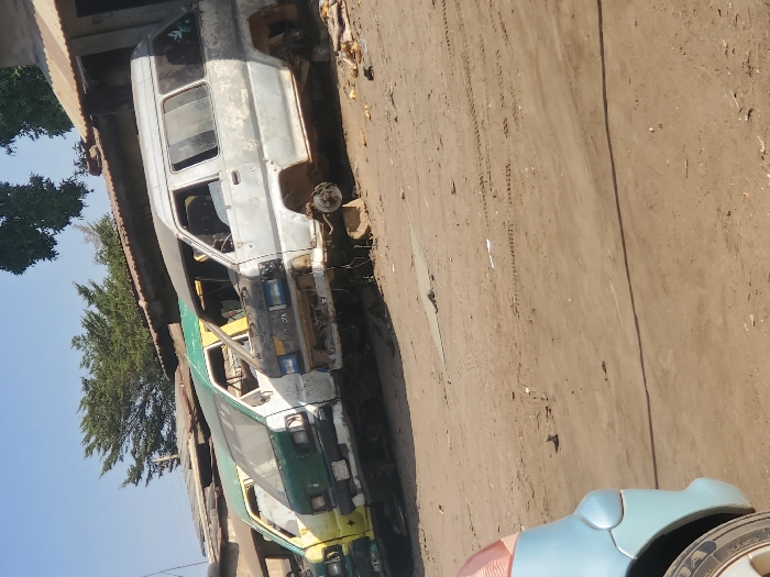 This image displays a vehicle salvage or long-term storage yard situated in Kaduna South, Nigeria, under bright daylight. The foreground reveals the partial left front fender and tire of a light blue vehicle. Dominating the midground are three abandoned passenger vans, parked in a linear arrangement on an unpaved, reddish-brown dirt lot exhibiting tire tracks and minor debris. From left to right, a yellow van and a green van are visible, both appearing dirty and worn. To their right, a heavily stripped gray/silver Toyota passenger van is present; its front bumper, headlights, and all four wheels are missing, and it rests on visible support blocks. The interior of this gray van is exposed and appears disassembled. In the background, low-rise structures with corrugated metal roofs (brown and green) are discernible, topped by sparse green trees against a clear blue sky. No individuals or specific actions are observed within the scene.