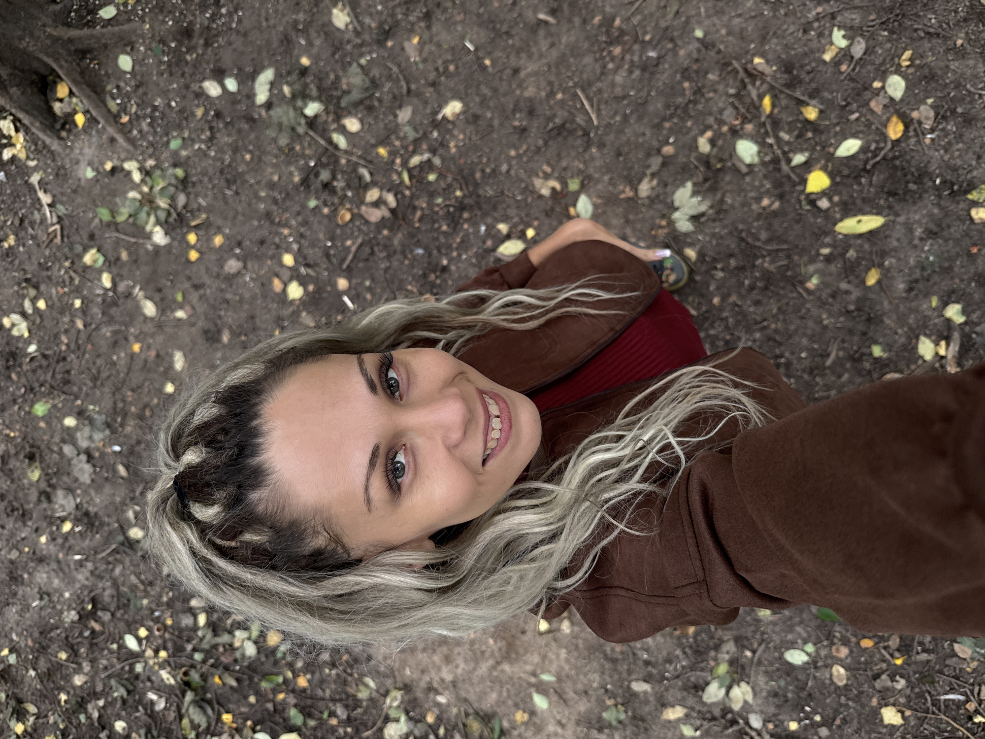 A high-angle, medium shot shows a woman with blonde hair styled in a ponytail, looking up and smiling directly at the camera. She is wearing a brown jacket over a red top. Her hair is a mix of blonde and darker roots, with some braided sections at the top. The background is a dirt path covered with fallen leaves and small branches, suggesting an outdoor, natural setting. There are no discernible city landmarks or architectural features present to indicate a specific location within Dumino, Russia. The overall impression is a casual, candid photograph taken outdoors.