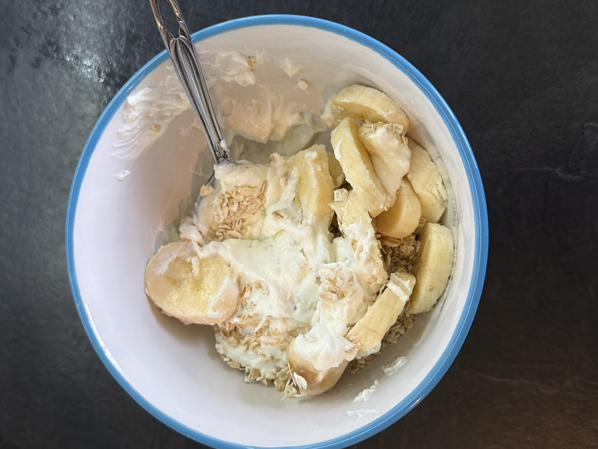An overhead view captures a white ceramic bowl with a light blue rim, partially filled with food. The contents consist of sliced yellow bananas, a creamy white substance resembling yogurt, and light brown rolled oats, all intermixed. A silver-colored metal spoon, featuring a decorative handle, is positioned within the food on the right side of the bowl. The bowl rests on a dark, textured surface, likely a countertop or table. The scene is an indoor domestic setting, reportedly situated in Pleven, Bulgaria. No individuals are present.