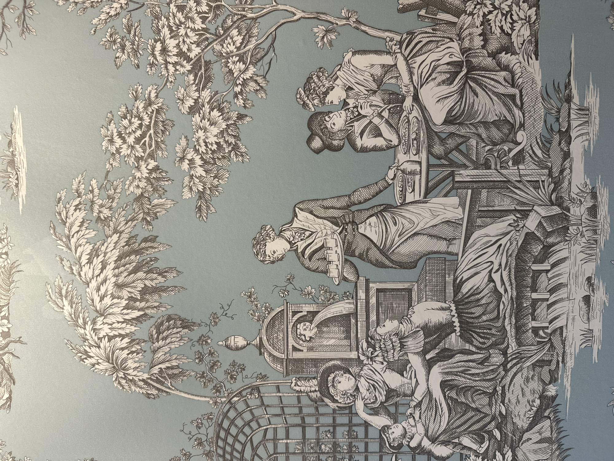 The image depicts a toile-style illustration on a light blue background. The scene portrays a gathering of people in an outdoor setting, possibly a garden or park. In the foreground, several individuals are seated, including a child on a woman's lap. A prominent fountain with a carved face spouts water. A server stands to the right, holding a tray of small items, likely food or drinks. Further to the right, a couple is seated at a table, engaged in conversation or sharing a flower. Trees with detailed foliage are scattered throughout the scene, creating a sense of an idyllic, possibly historical, outdoor environment. The illustration is rendered in black and white, with a focus on intricate linework and shading. There is no discernible indication or clue within the image that places the scene in Minsk, Belarus.